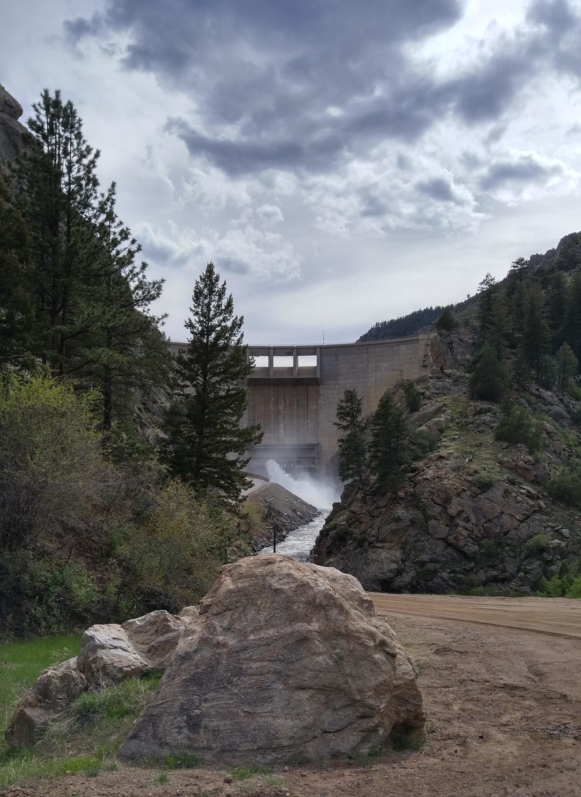 A concrete dam surrounded by rocky hills and tall pine trees. Water is being released from the dam, cascading down a narrow channel. The sky is partly cloudy, with a mix of gray and blue tones. A large rock in the foreground and a dirt path lead up to the scene. Waterton Canyon mountain bike trail.