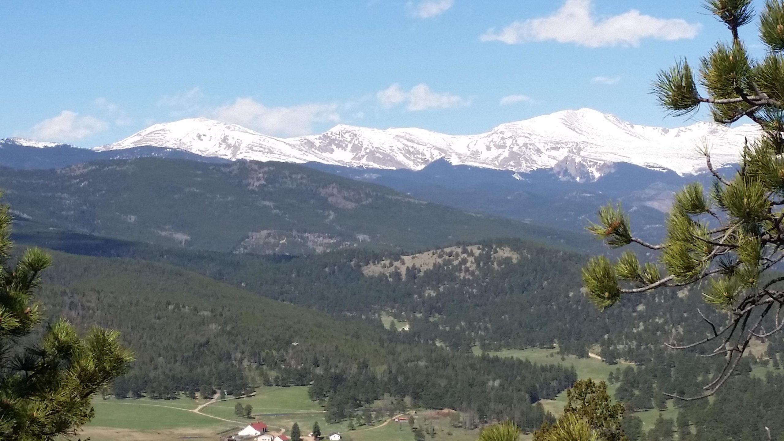 A scenic view of snow-capped mountains in the distance, surrounded by lush green valleys and forests. A few small buildings with red roofs are visible in the foreground, set among the rolling hills. The sky is clear with a few scattered clouds. 3 Sisters / Alderfer mountain bike trail.