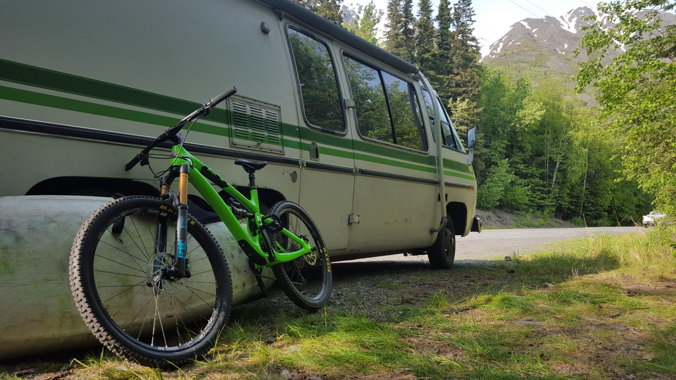 Yeti SB6: A bright green mountain bike is propped against a white van with green stripes, set in a lush, green landscape with trees and mountains in the background. The scene captures a sense of adventure and outdoor exploration.