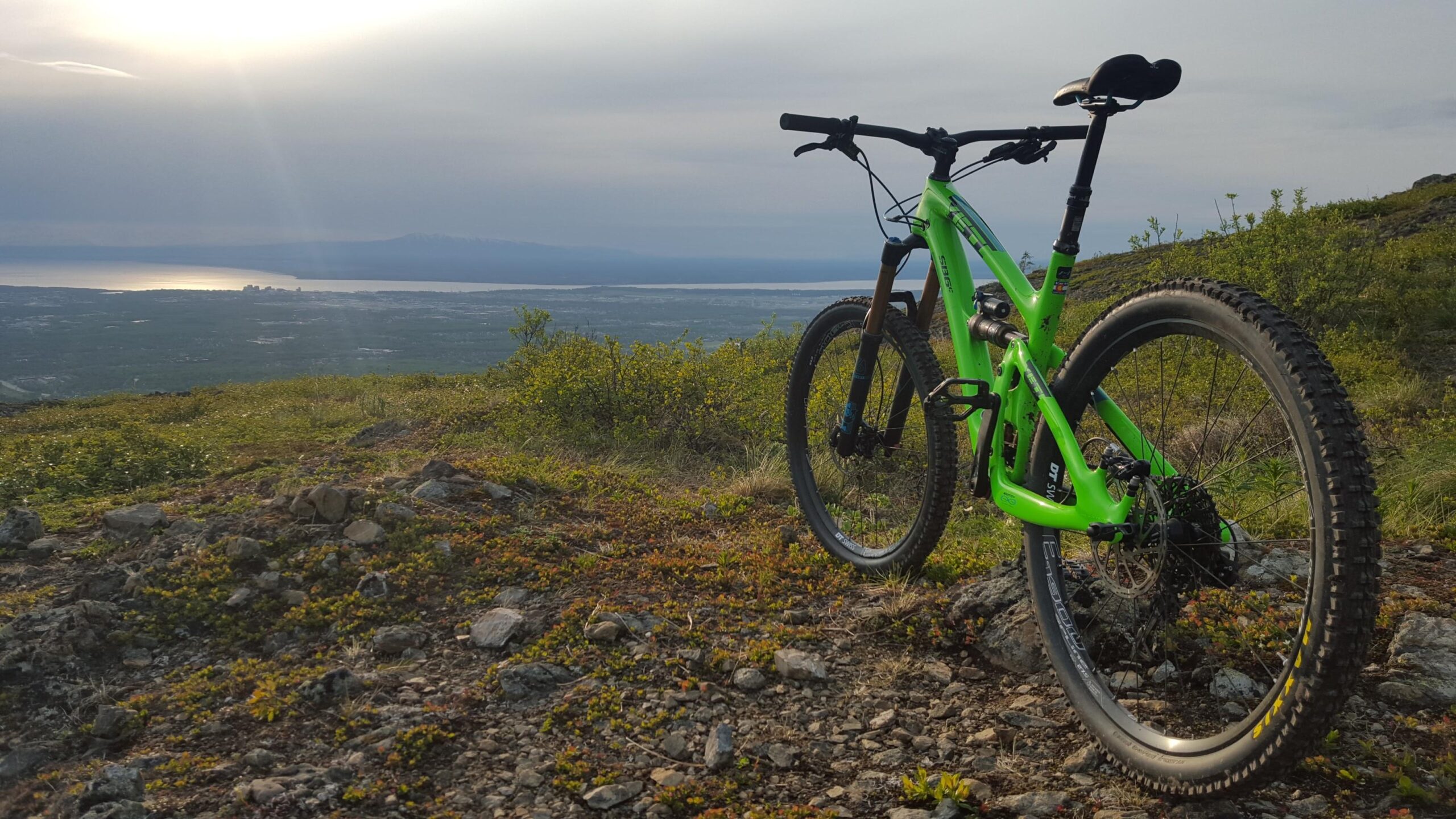 Yeti SB6: A vibrant green mountain bike stands on rocky terrain, overlooking a vast landscape with a body of water in the distance under a cloudy sky. The sun's rays create a shimmering effect on the water, while patches of greenery and wildflowers surround the bike, adding to the scenic outdoor setting.