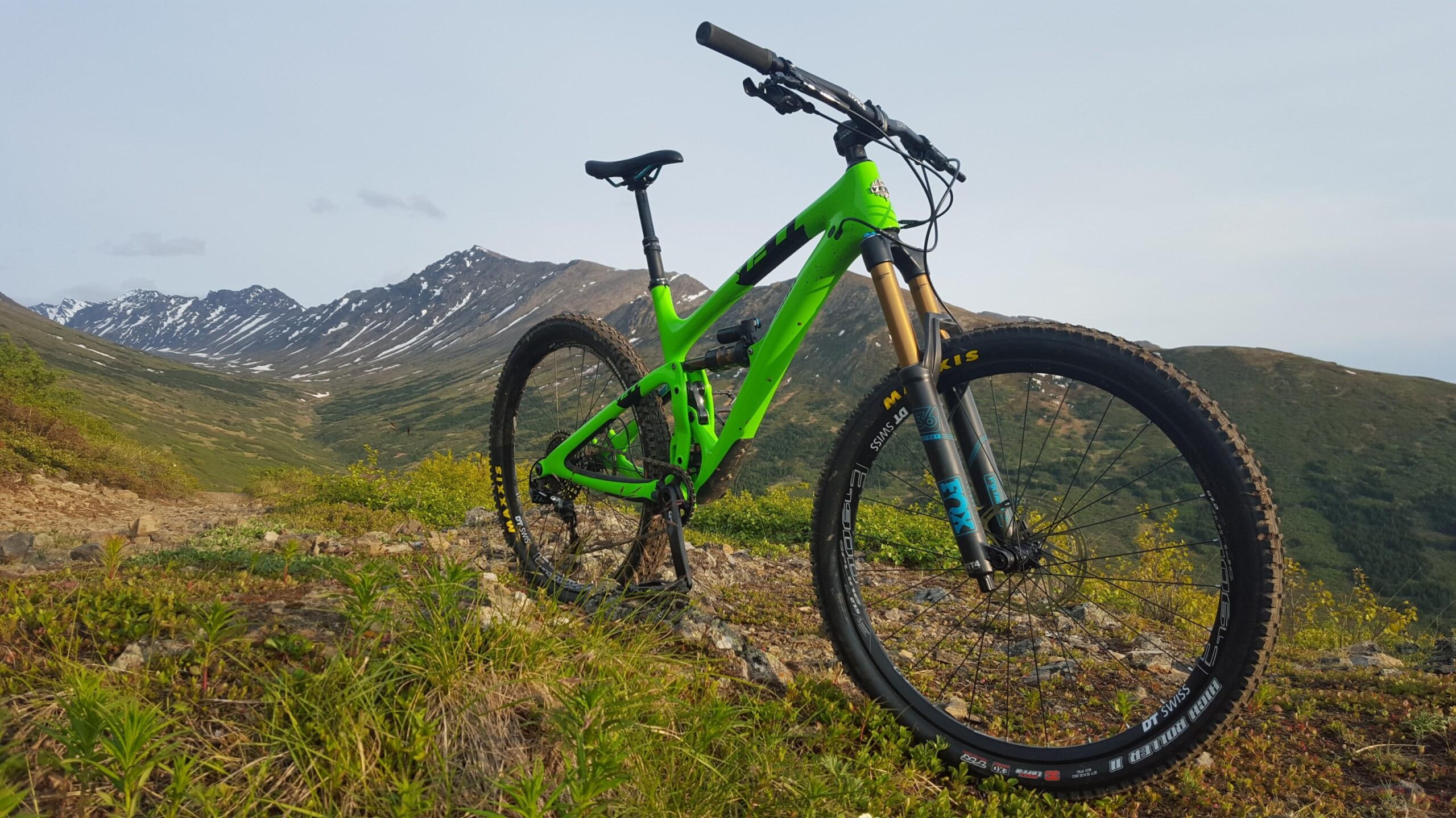 Yeti SB6: A vibrant green mountain bike is positioned on rocky terrain, with scenic mountains and valleys in the background. The setting features lush greenery and patches of snow on the distant peaks, under a clear sky.