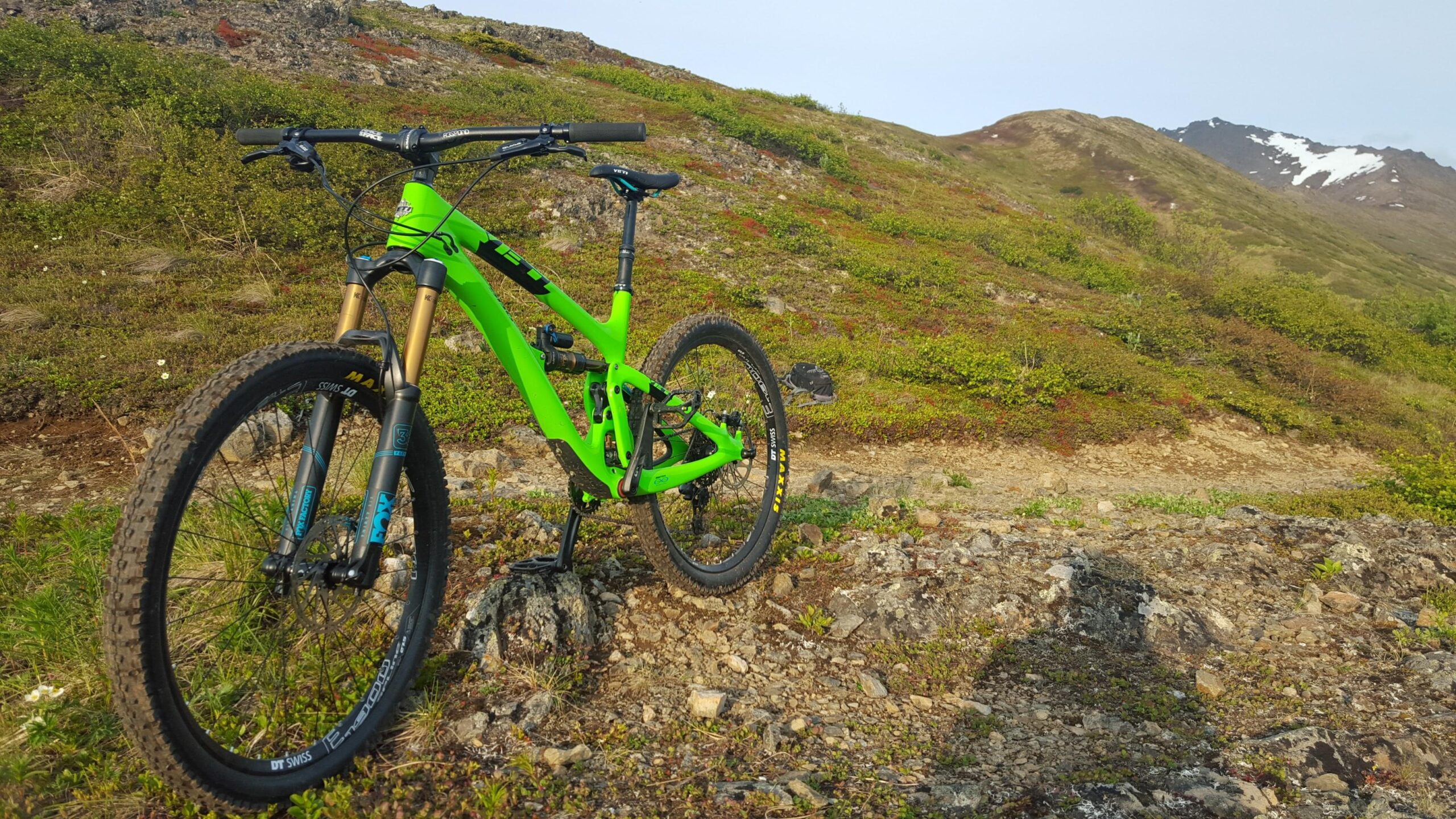 Yeti SB6: A bright green mountain bike is parked on rocky terrain, surrounded by lush greenery and distant hills under a clear sky. The bike's front wheel and suspension are prominently displayed, showcasing its rugged design, while the backdrop features a natural landscape with a slight incline and some patches of snow on the mountain tops.