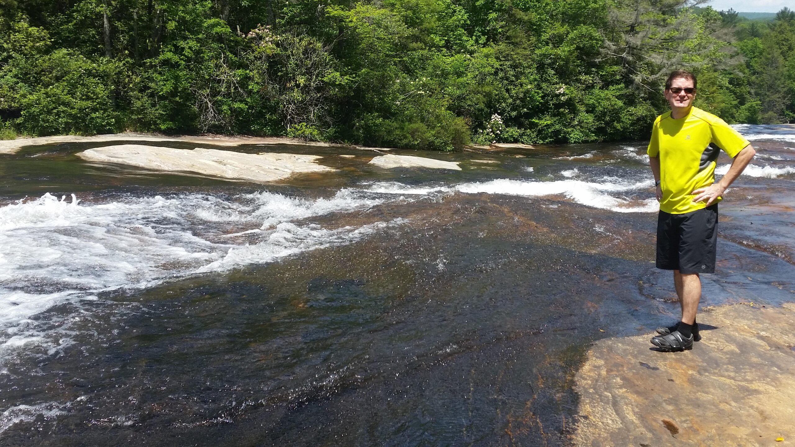 A person standing on a rocky riverbank, smiling while wearing a bright yellow shirt and black shorts. The river is flowing nearby, with white water rapids and lush greenery in the background under a clear blue sky. Mine Mountain mountain bike trail.