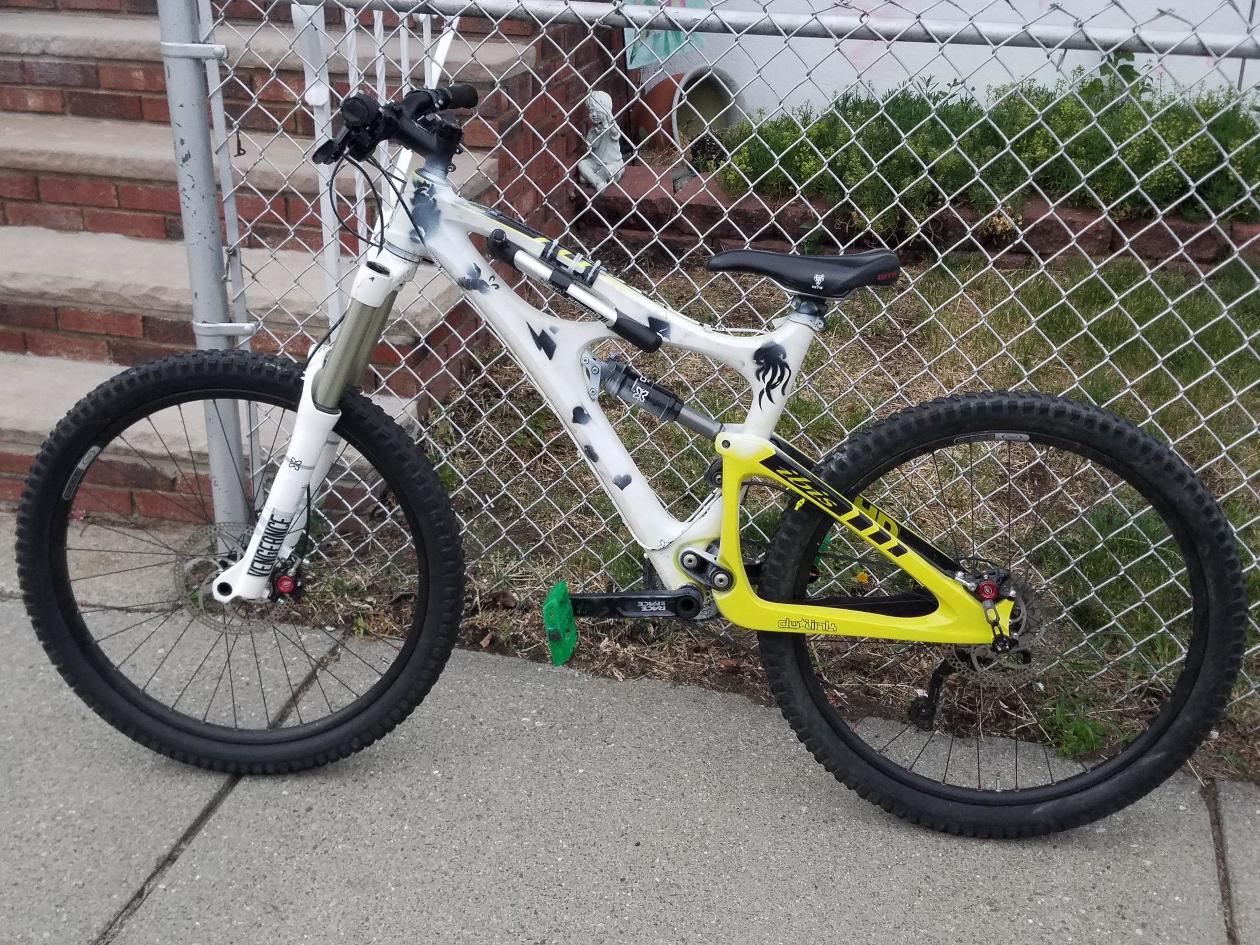 A mountain bike with a white frame featuring black and yellow accents, parked near a chain-link fence. The bike has chunky tires suited for rough terrain and is positioned on a concrete surface, with greenery visible in the background. Lynn Woods Reservation mountain bike trail.