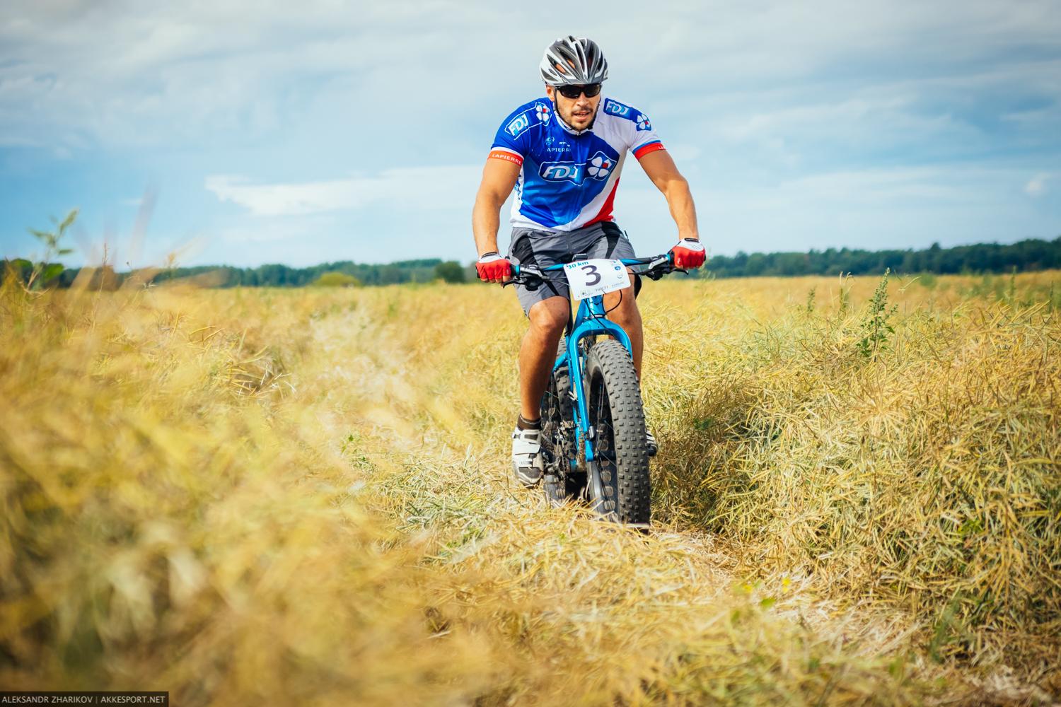 Felt Double Double: A cyclist wearing a blue and white jersey rides a mountain bike through a field of tall, dry grass under a blue sky with scattered clouds. The bike features thick tires suitable for rugged terrain, and the cyclist has a race number attached to their jersey.