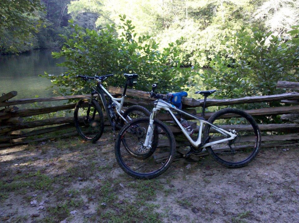 Trek Superfly: Two mountain bikes are parked near a rustic wooden fence by a serene body of water, surrounded by lush greenery. The scene captures a peaceful outdoor setting, ideal for biking enthusiasts.