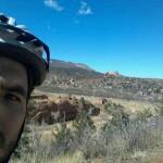 A person wearing a bike helmet stands in a natural landscape with a blue sky in the background. The scenery features rolling hills, sparse vegetation, and distant rock formations. Red Rock Canyon mountain bike trail.