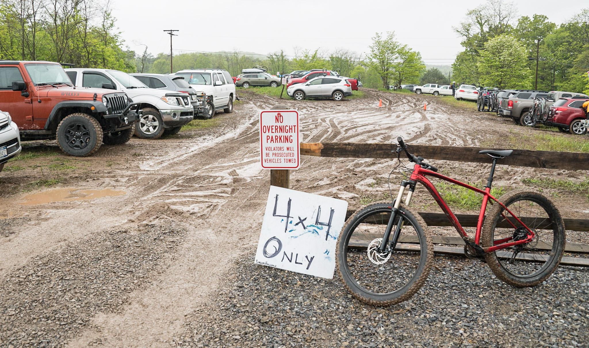 Specialized Fuse Comp 6Fattie: A muddy parking area filled with various vehicles, including SUVs and trucks, with a red bicycle parked against a wooden fence. A sign indicating "No Overnight Parking" and another sign that reads "4x4 Only" are visible in the foreground. The scene is set in a green, wooded area, and it appears to be rainy or wet.