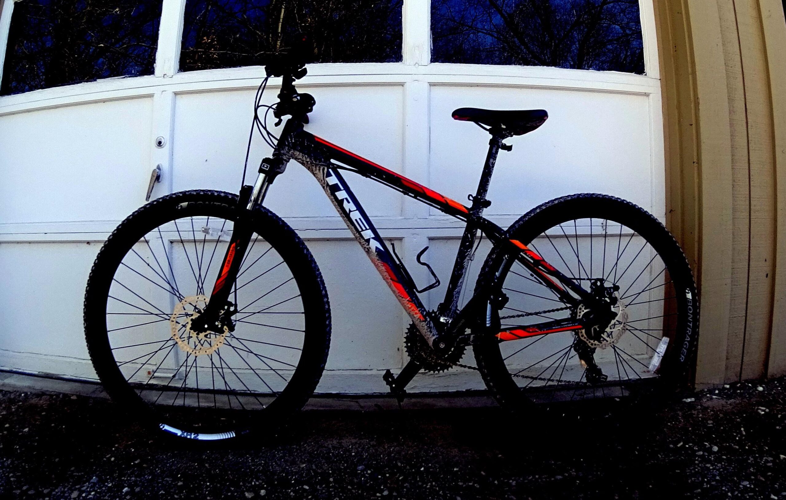 Trek Marlin 5: A mountain bike leaning against a garage door, with a black and orange frame and knobby tires. The bike appears slightly dirty, indicating recent use on rugged terrain.