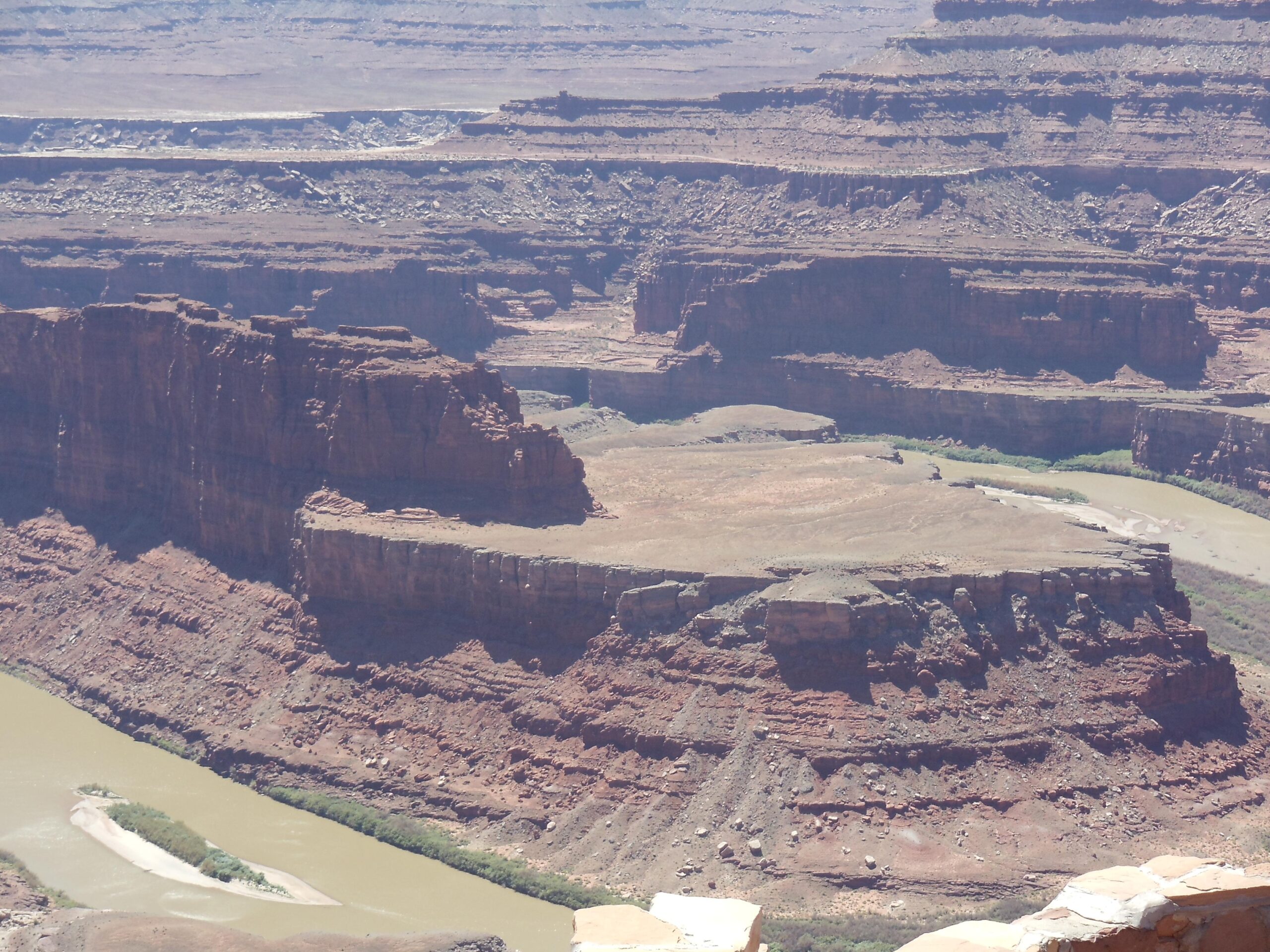 A panoramic view of a rugged canyon landscape featuring steep red rock formations and a winding river below, surrounded by dry terrain and distant cliffs under a clear blue sky. Dead Horse Point State Park mountain bike trail.