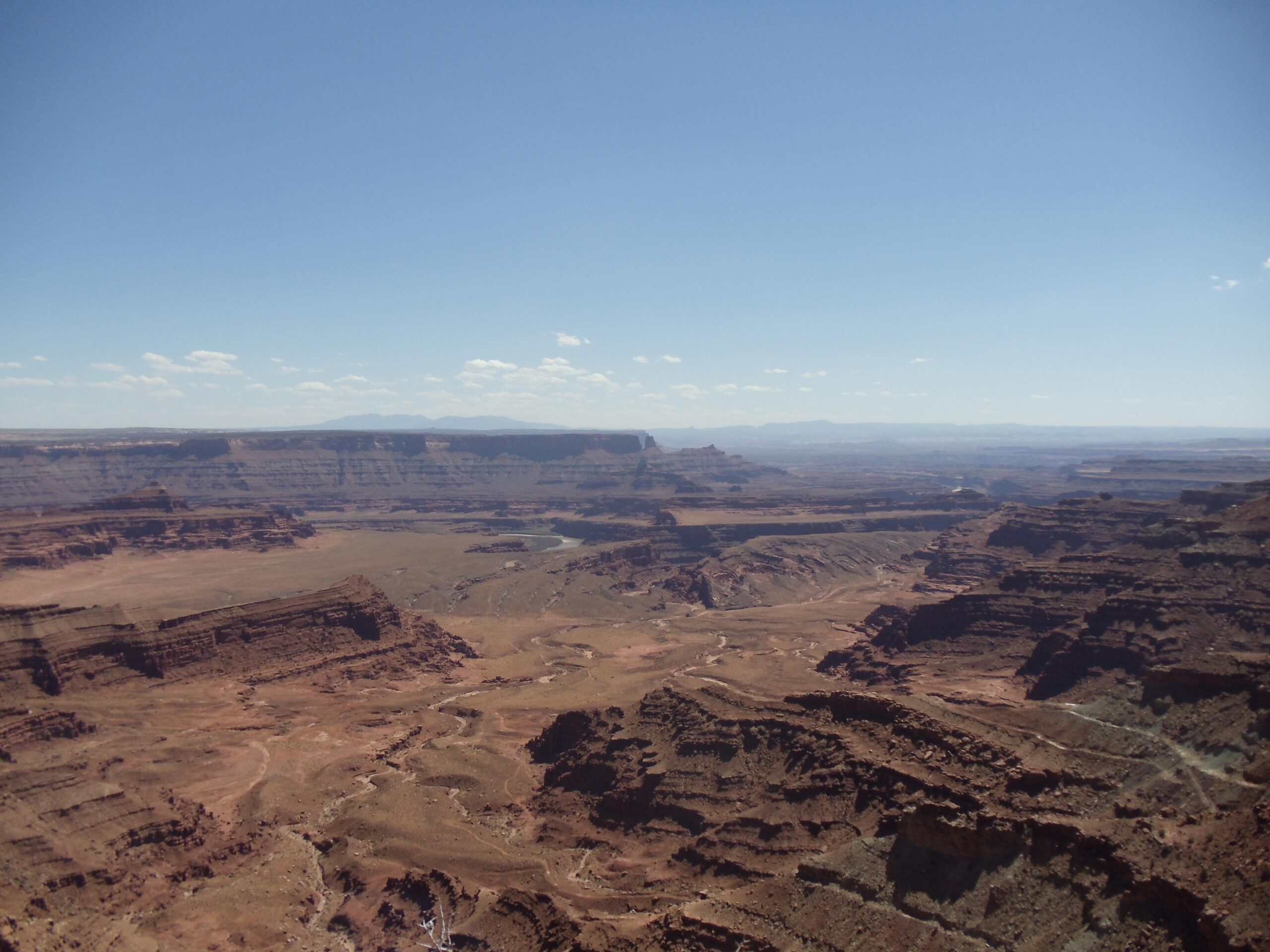 A wide view of a rugged canyon landscape featuring layered rock formations, with a clear blue sky and scattered clouds above. The terrain includes deep valleys and winding roads, showcasing the natural beauty of the arid environment. Dead Horse Point State Park mountain bike trail.