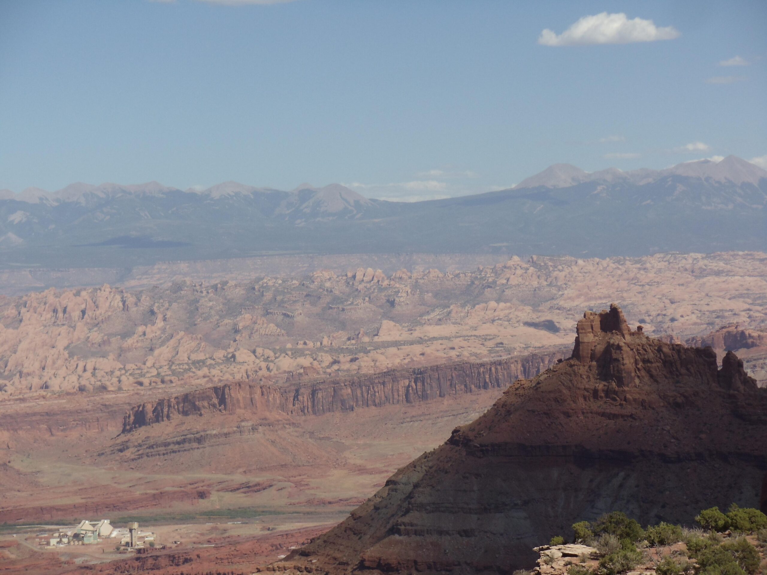 A panoramic view of a rugged desert landscape featuring layered rock formations, distant mountains, and a small building in the valley below. The sky is clear with a few clouds, highlighting the vastness of the scene. Dead Horse Point State Park mountain bike trail.