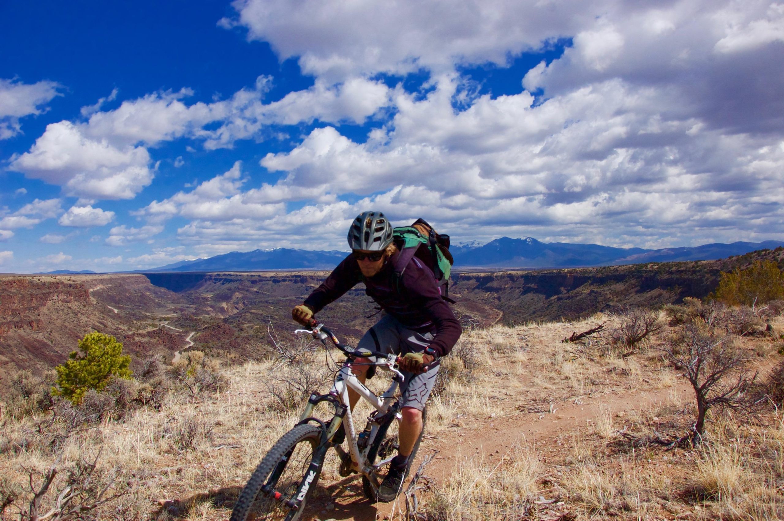 A mountain biker navigates a rugged trail in a scenic canyon landscape, surrounded by dry vegetation and distant mountains under a partly cloudy blue sky. Taos Valley Overlook mountain bike trail.
