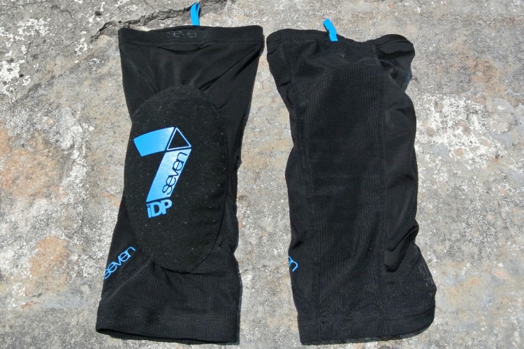 A pair of black protective elbow sleeves, featuring padded sections and blue branding. The sleeves are displayed on a textured, light-colored surface.
