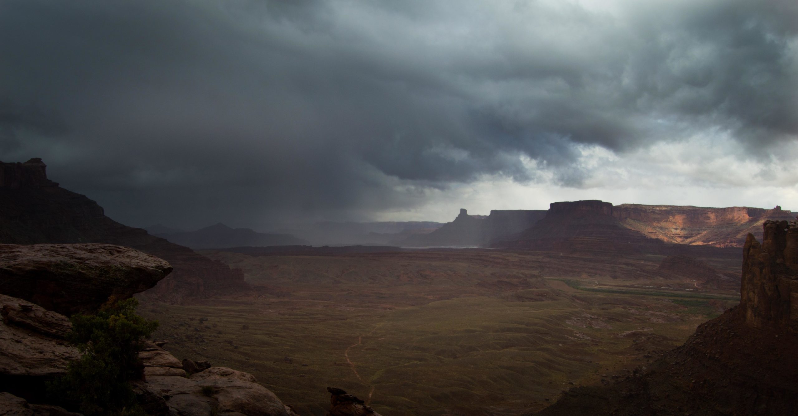 A scenic landscape featuring dramatic, dark storm clouds overhead, with rugged red rock formations and an expansive valley below. A winding path is visible through the grassy terrain, and distant cliffs rise sharply in the background, illuminated by patches of light breaking through the clouds. Hymasa mountain bike trail.
