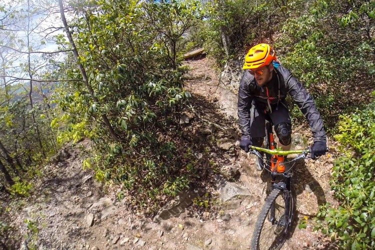 A mountain biker navigates a rocky trail surrounded by greenery, wearing a bright orange helmet and black riding gear. The cyclist appears focused as they maneuver their bike over the uneven terrain under a partly cloudy sky.