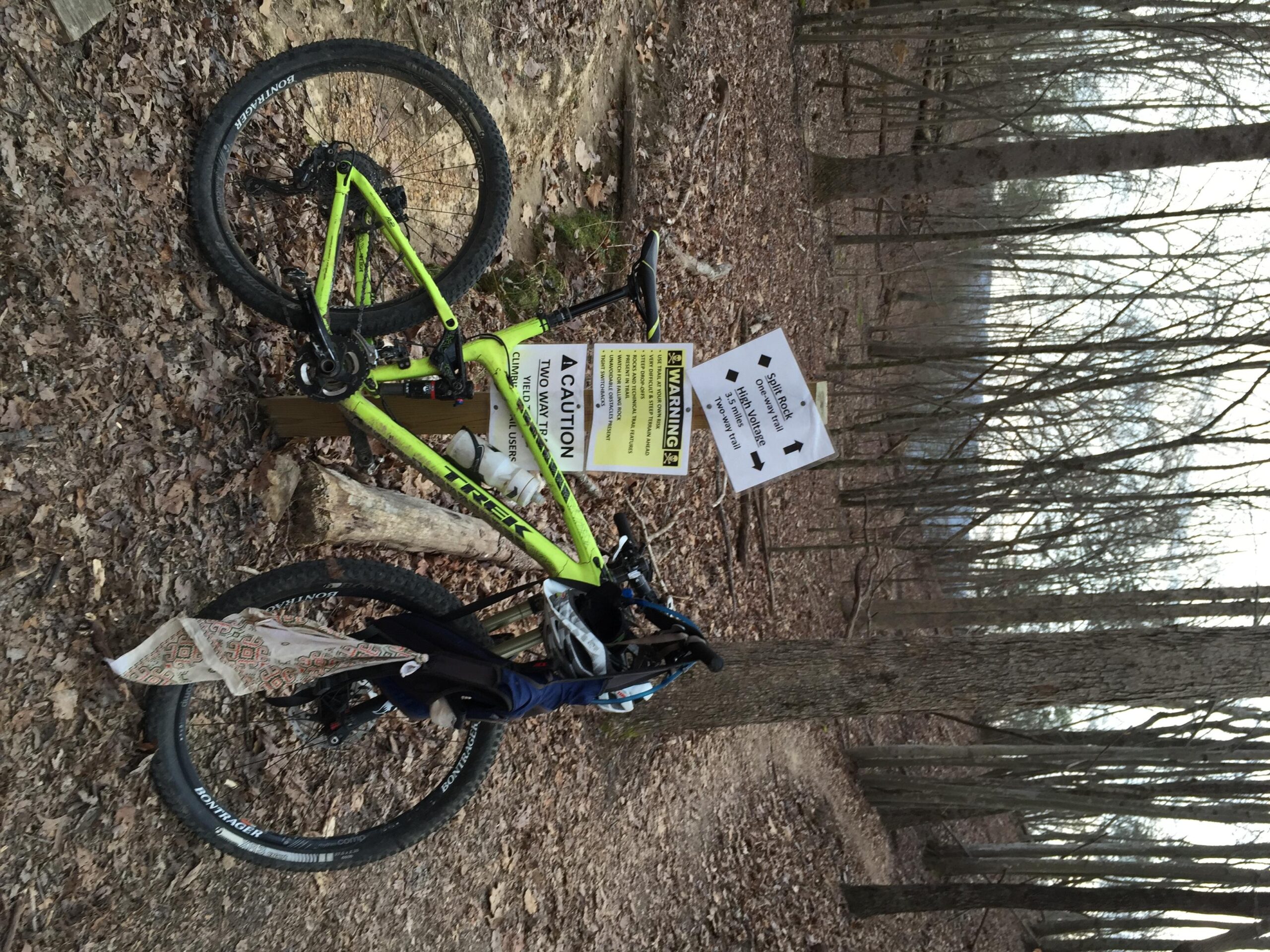 Trek Trek Fuel EX 9.8 27.5: A green mountain bike resting against a tree in a wooded area, surrounded by fallen leaves. A caution sign is attached to the bike, with another sign nearby indicating directions for trail use. The background features bare trees, suggesting a winter or early spring setting.