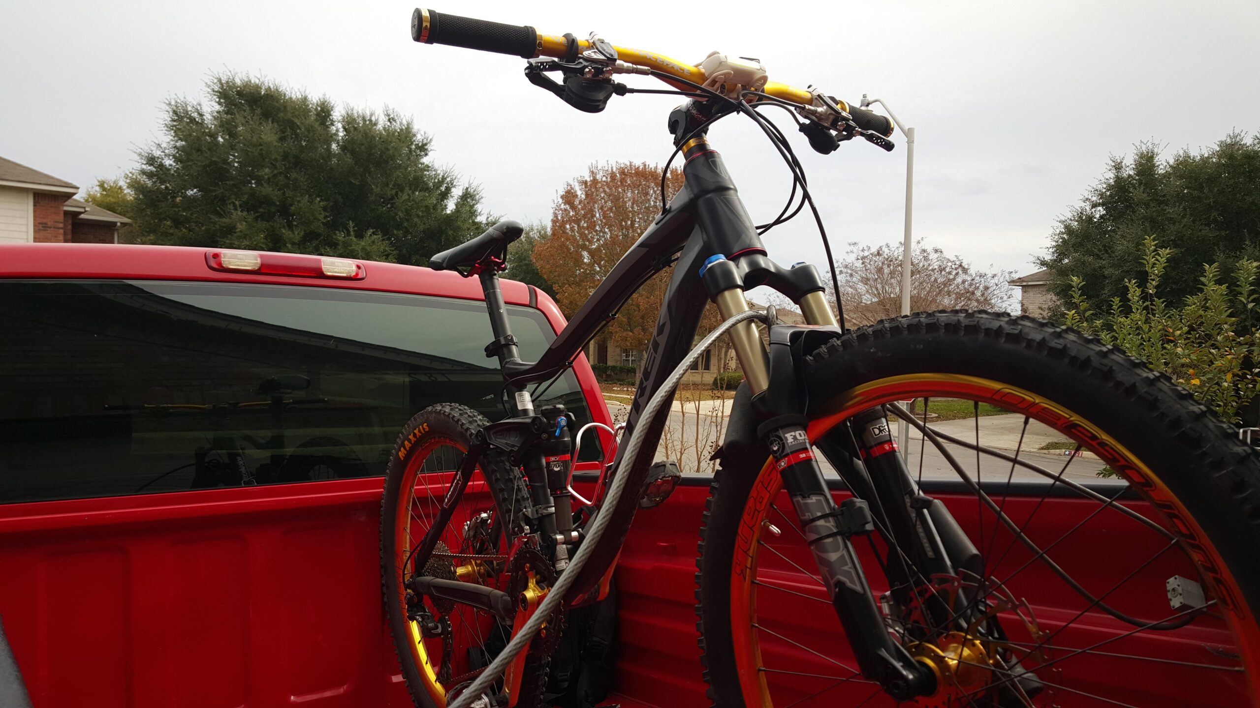 Trek Fuel EX 8: A mountain bike with a black frame and bright yellow handlebars is secured in the back of a red pickup truck. The bike features thick tires and front suspension forks, with a slightly cloudy sky in the background and residential buildings visible.
