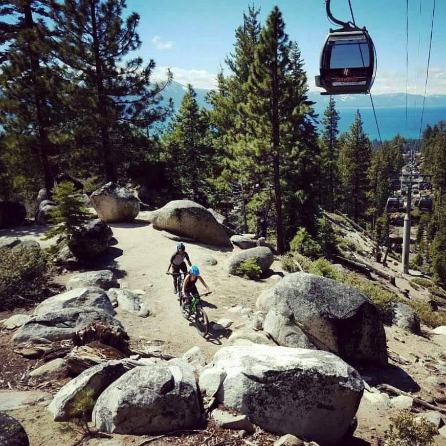Two mountain bikers riding on a gravel path surrounded by large rocks and pine trees, with a scenic view of a lake and distant mountains under a clear blue sky. A gondola lift is visible in the background. Tahoe Rim Trail mountain bike trail.