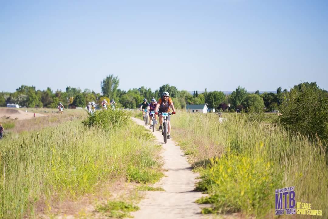 A group of mountain bikers riding along a dirt path surrounded by tall grass, under a clear blue sky. The scene captures the excitement of an outdoor biking event with several participants visible in the background. Ada/Eagle Bike Park mountain bike trail.