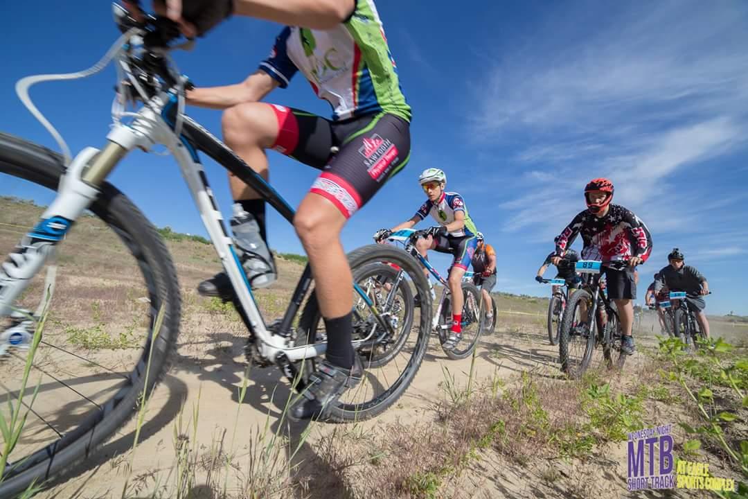 A group of mountain bikers riding on a dirt trail under a blue sky. They wear colorful cycling jerseys and helmets, showcasing a variety of bicycles as they navigate through a natural landscape. The scene captures the dynamic movement of the riders, highlighting their determination and teamwork during a biking event. Ada/Eagle Bike Park mountain bike trail.