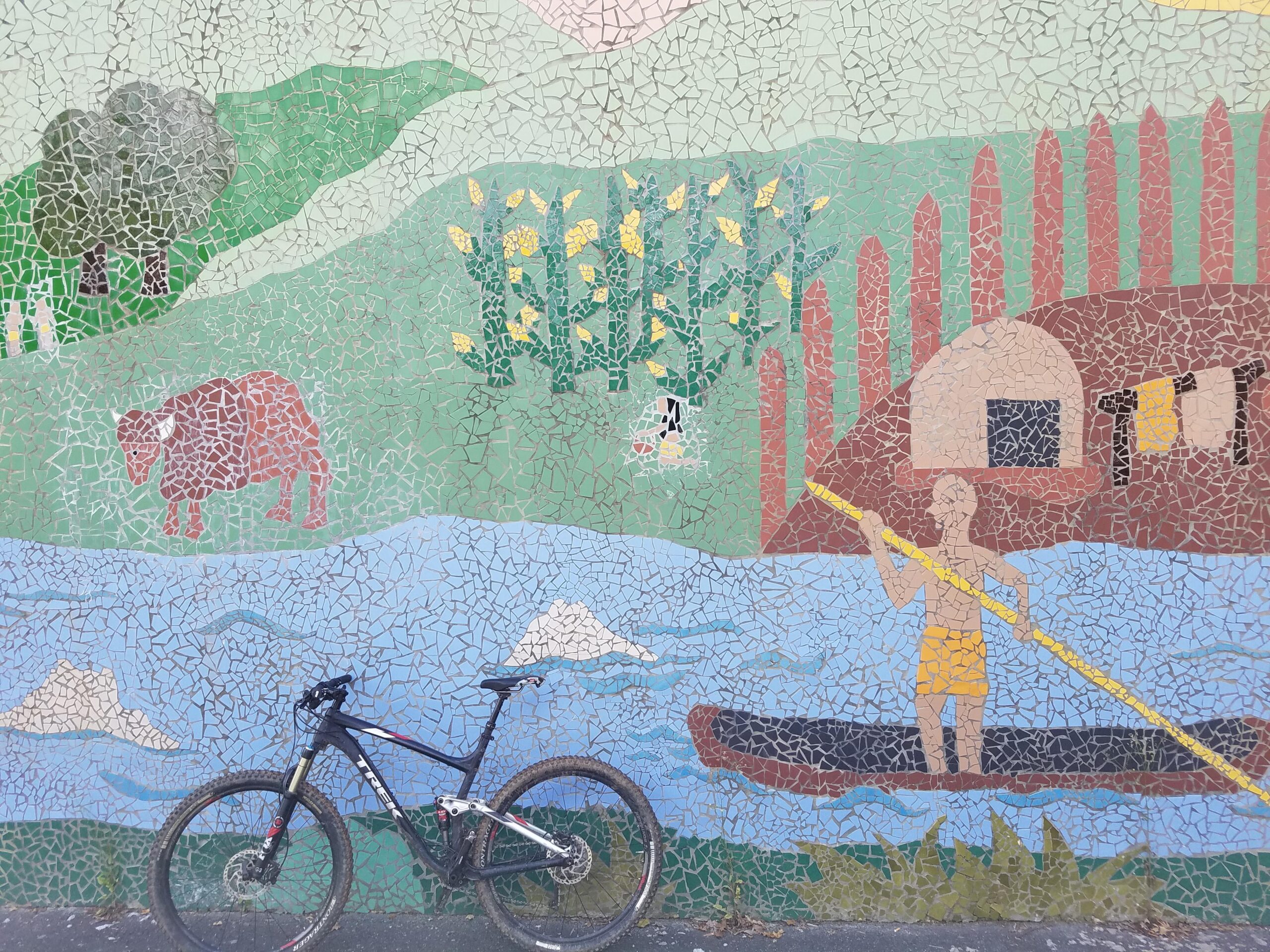 Trek Fuel EX 8: A colorful mosaic mural depicting a rural scene with a cow grazing, a person paddling a canoe on a river, and a simple house. The background features green hills, trees, and corn plants. In the foreground, a bicycle leans against the wall.