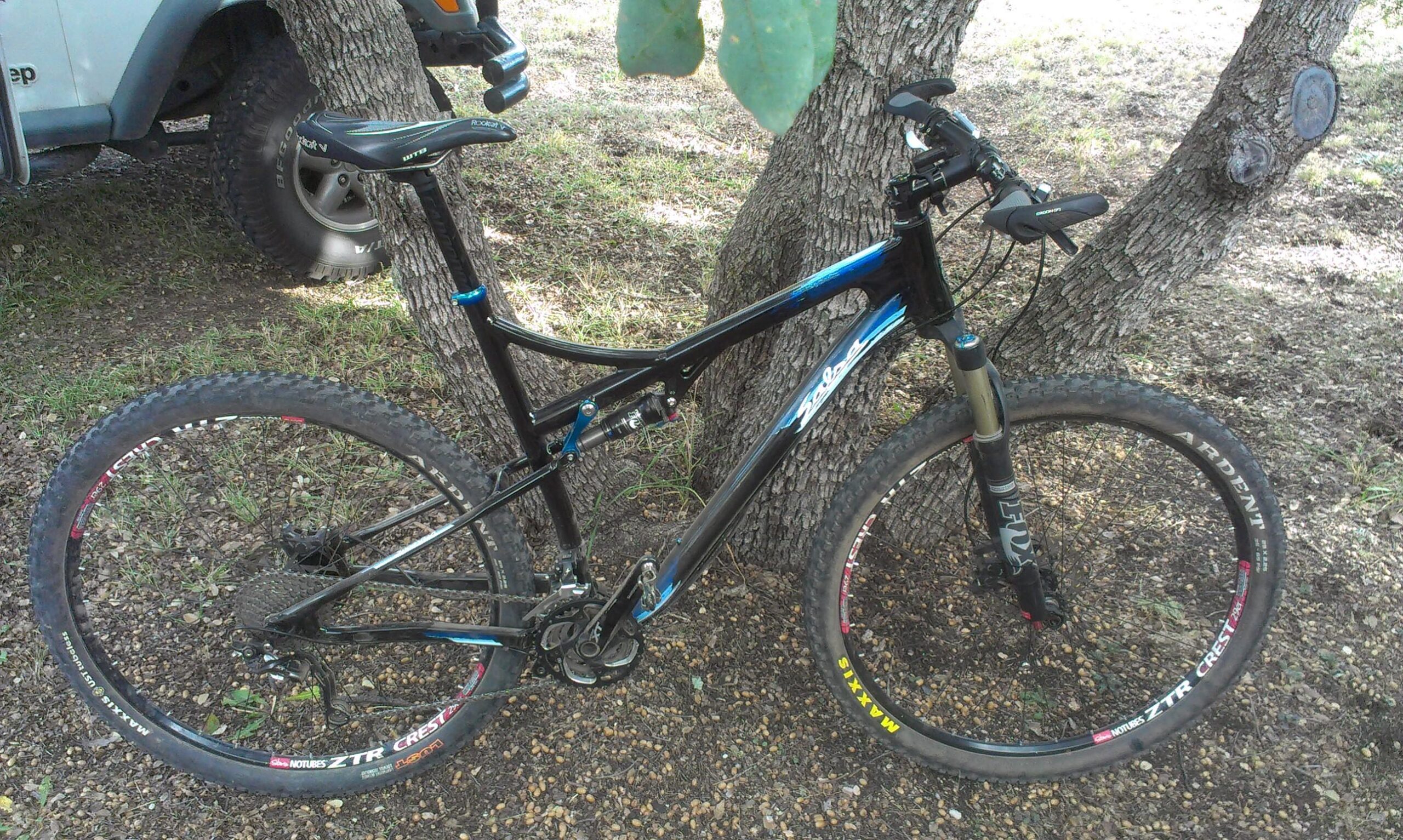 Salsa Spearfish: A black mountain bike with blue accents leaning against a tree. The bike has knobby tires and a suspension system, parked on a gravel surface under a tree. A partially visible blue Jeep is in the background.