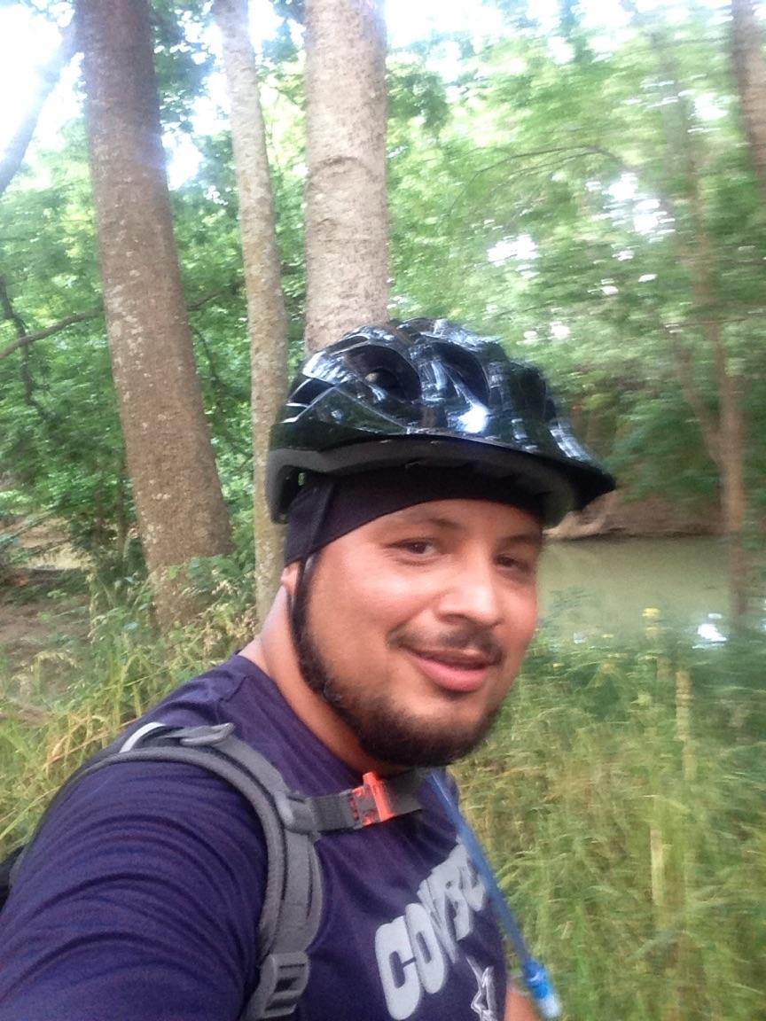A person wearing a black helmet and a blue t-shirt stands in a forested area, smiling at the camera. The background features tall trees and greenery, with a glimpse of water visible. The individual is equipped with a backpack and hydration pack. Rowlett Creek Preserve mountain bike trail.