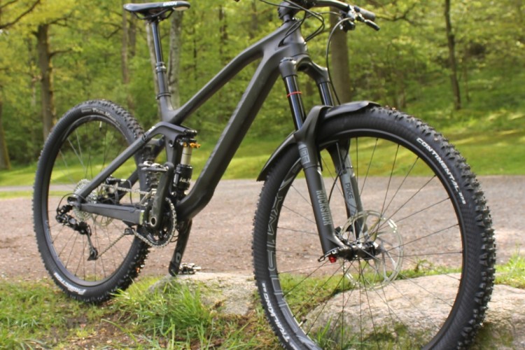 A sleek black mountain bike positioned on a grassy area near a rocky path. The bike features wide tires, a sturdy frame, and a high-performance suspension system, surrounded by lush greenery in the background.