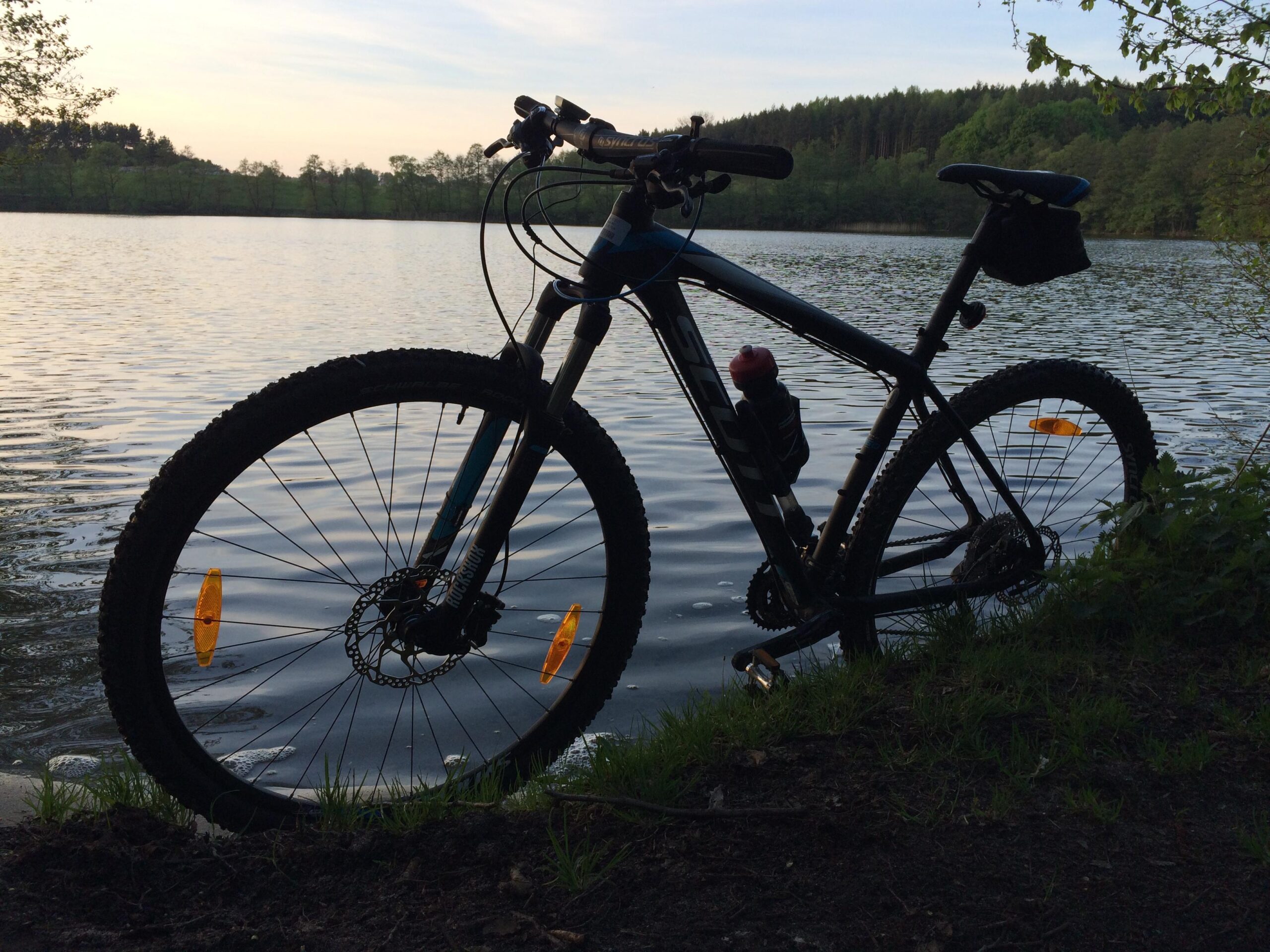 Scott Scale 920: A mountain bike parked near the edge of a calm lake, with the water reflecting the sky. The bike is partially in the foreground, showcasing its thick tires and a water bottle mounted on the frame. Surrounding the lake are trees and a grassy bank, creating a serene natural atmosphere. The time of day appears to be early evening, with soft lighting and a tranquil backdrop.