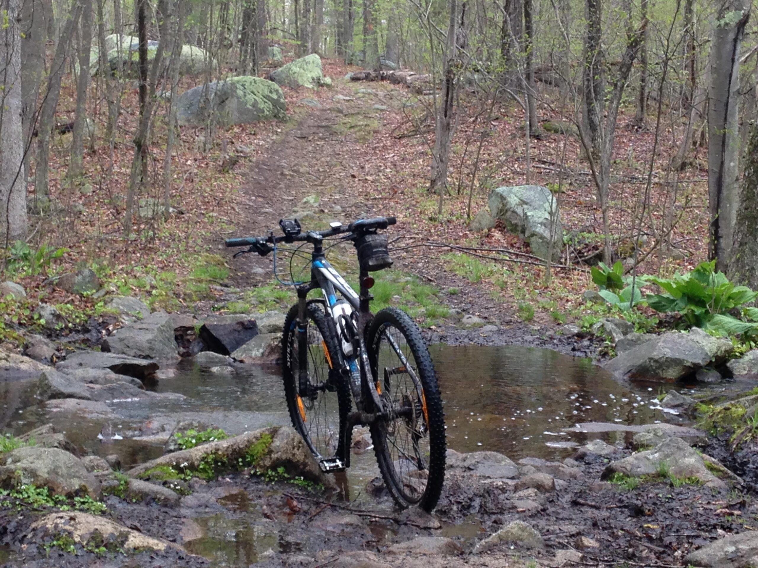 A mountain bike parked beside a small stream in a wooded area, with a dirt trail visible in the background. The ground is rocky and muddy, and there are scattered rocks and green foliage around the scene, indicating a natural outdoor setting. Vietnam Trails mountain bike trail.