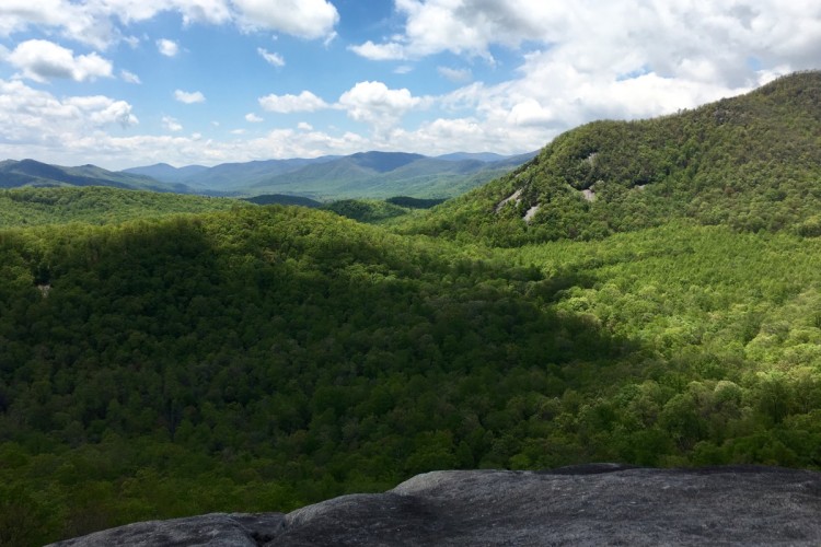 A panoramic view of lush green mountains under a partly cloudy sky. The foreground features a rocky ledge, with verdant hills and valleys stretching into the distance, revealing layers of rolling mountains and blue sky.