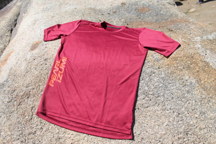 A maroon short-sleeve athletic t-shirt with the text "Pearl Izumi" printed in a lighter shade on the left side, laid flat on a rocky surface under natural lighting.