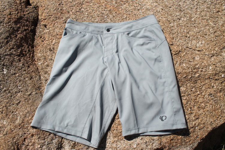 A pair of gray outdoor shorts laid flat on a textured rock surface, featuring a button closure and multiple pockets. The brand logo is visible on the hem.