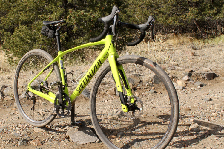 A bright yellow Specialized bicycle parked on a rocky dirt trail surrounded by trees and dry grass. The bike features drop handlebars, wide tires, and a small bag attached to the seat.