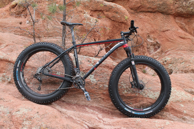 A black and red fat bike is positioned on rocky terrain, showcasing its wide tires and rugged frame. The backdrop features reddish rock formations and sparse vegetation, emphasizing the bike's suitability for off-road cycling.