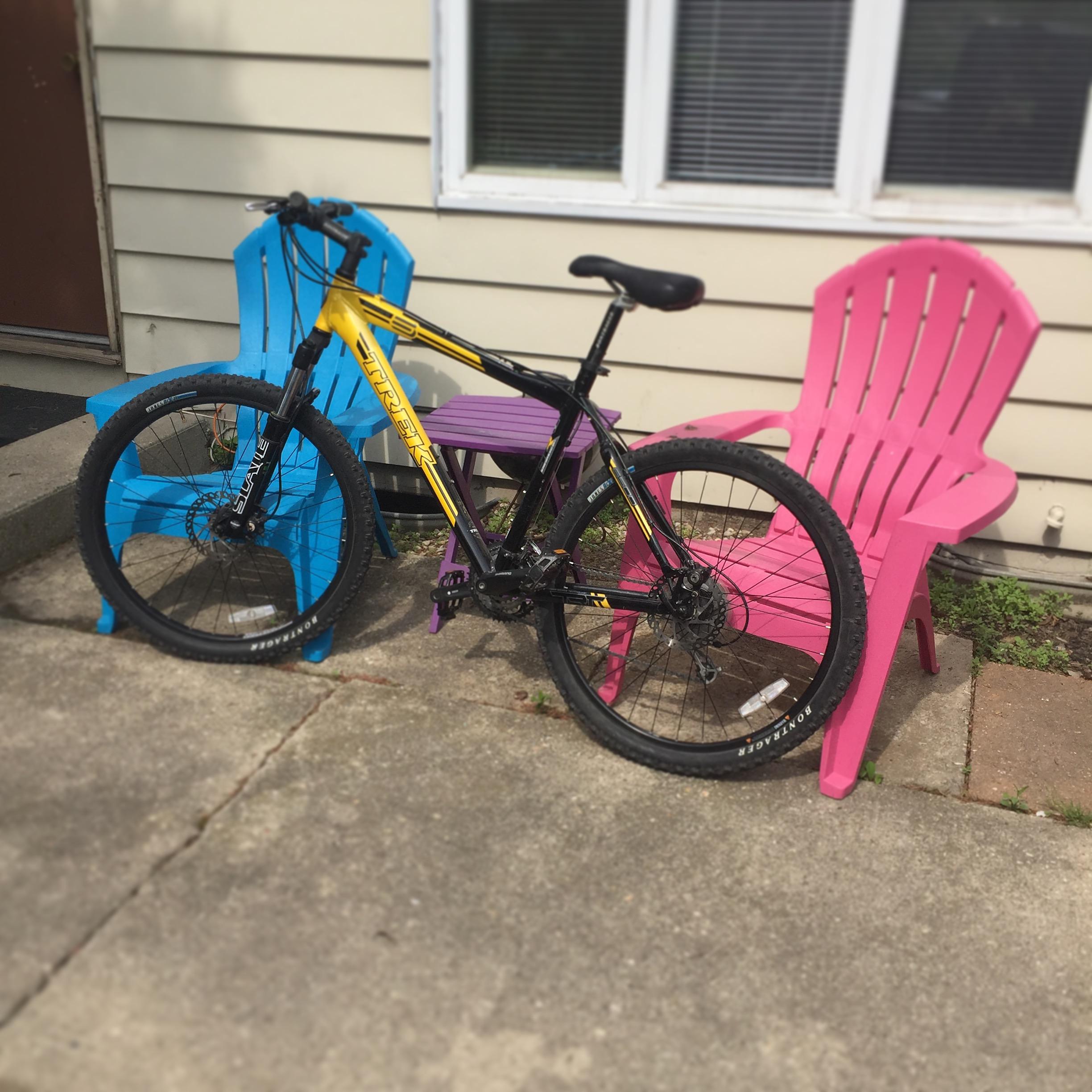 Trek 6500: A black mountain bike is leaning against vibrant pink and blue adirondack chairs on a concrete patio, with a beige house wall and window in the background.