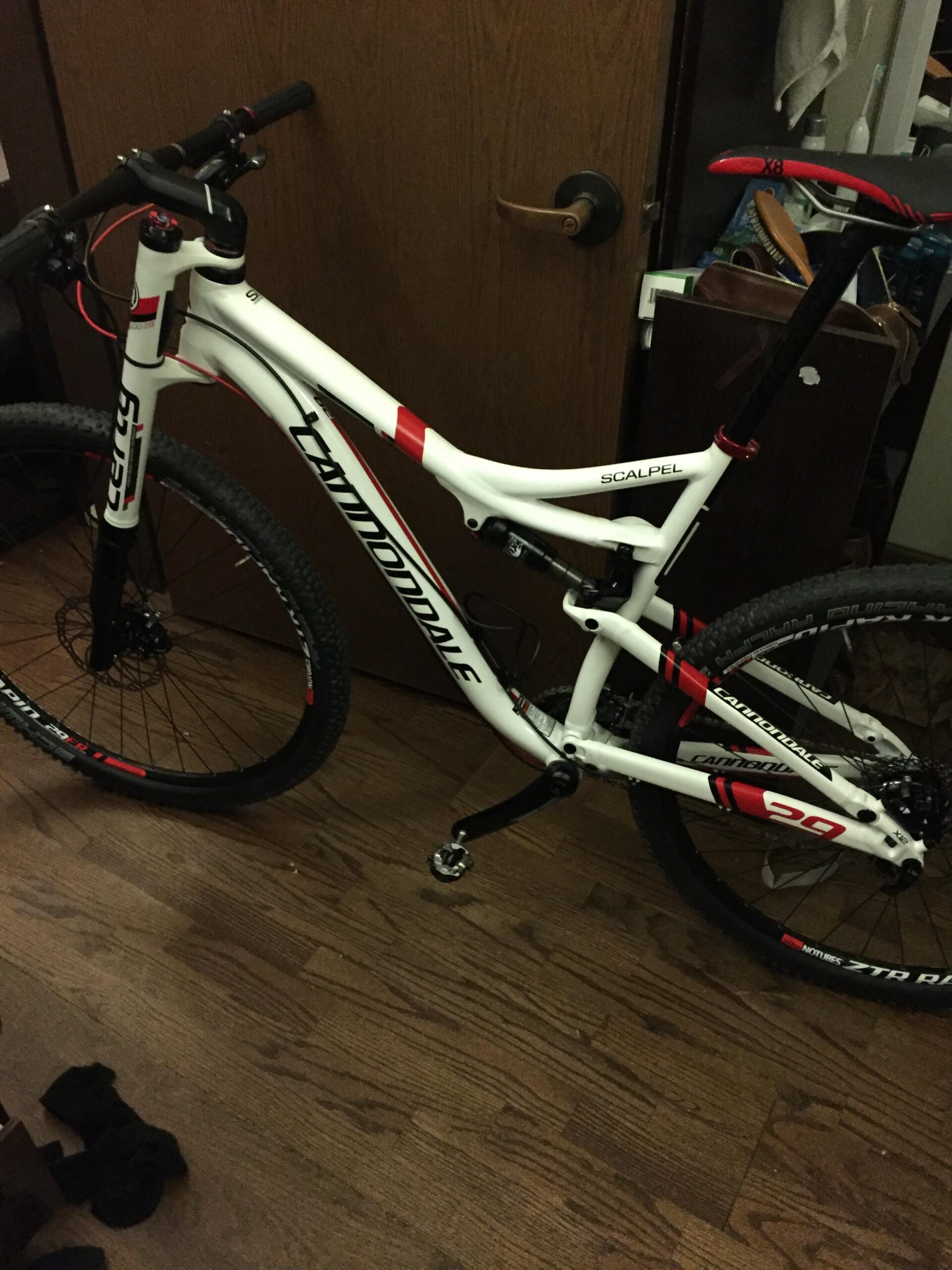 Cannondale Scalpel 29er 3: A white Cannondale Scalpel mountain bike leaning against a wooden door, featuring red and black accents. The bike has a dual-suspension design, wide tires, and is positioned on a wooden floor with some dark socks visible in the foreground.