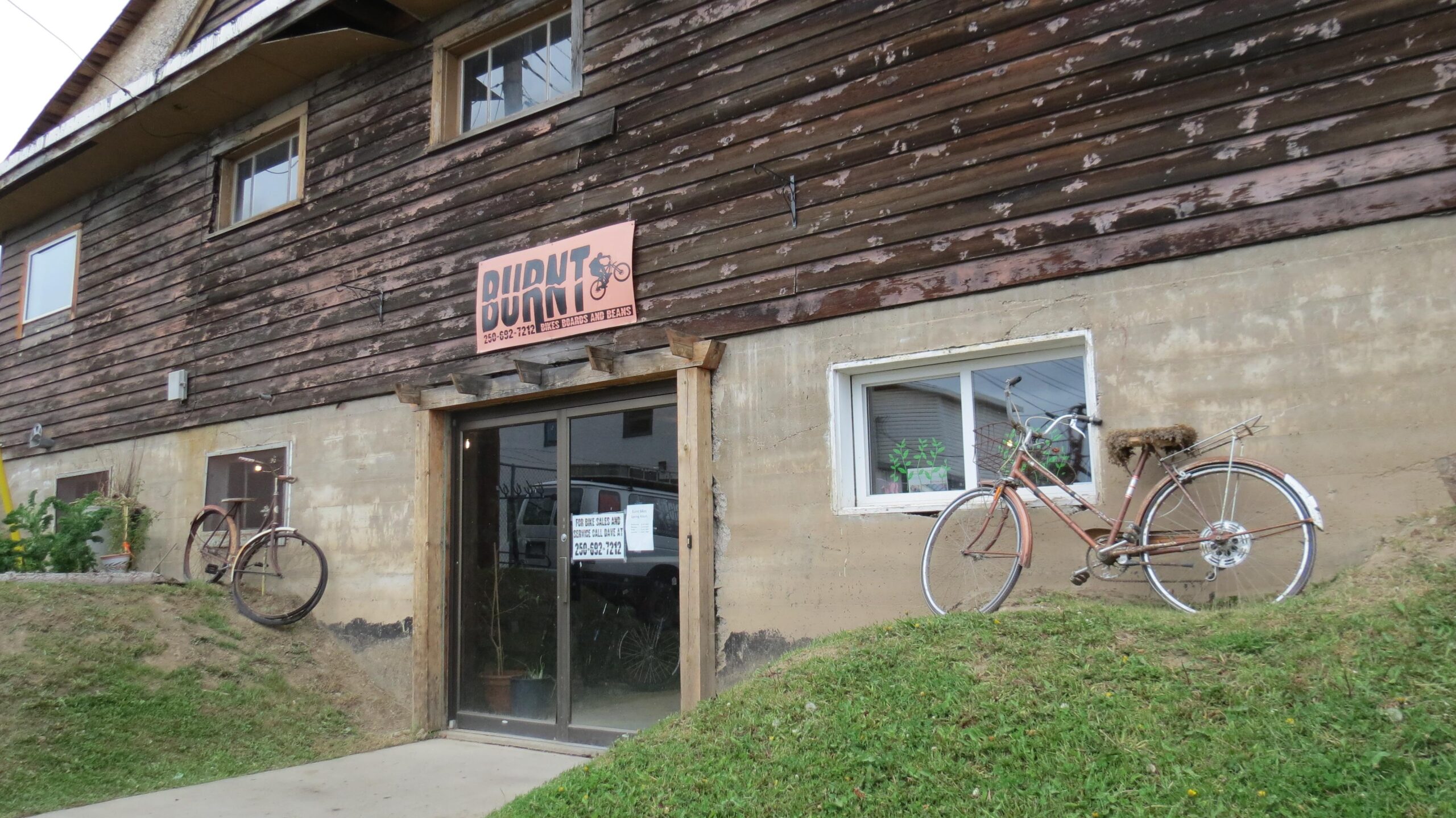 A rustic wooden building with a sign that reads "BURNT" is shown. The entrance features sliding glass doors and a window with a view of green plants inside. On either side of the entrance, there are bicycles mounted on the wall. The grassy area in front is slightly sloped, complementing the building's unique exterior.