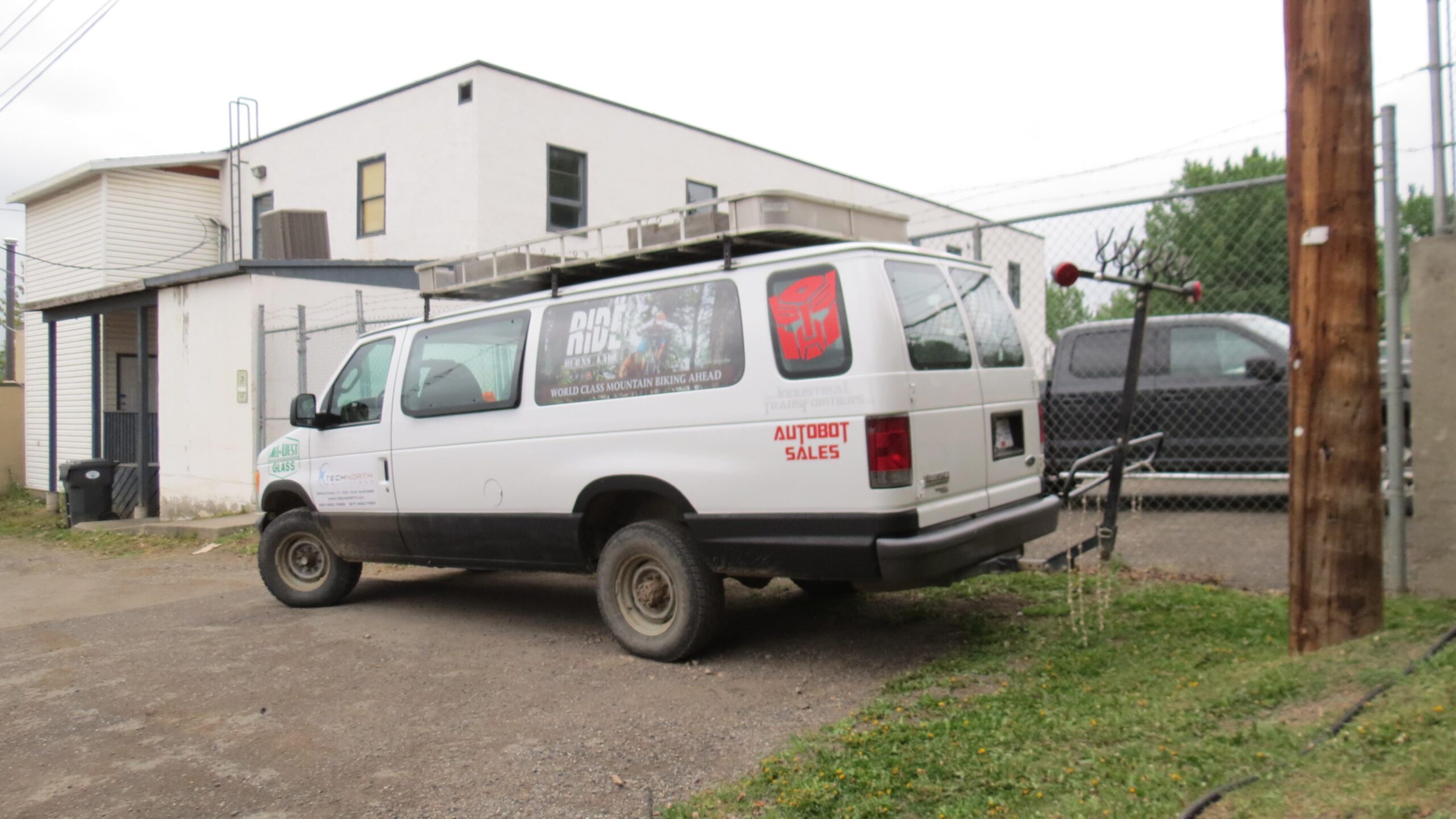 A white van parked in an alleyway, featuring advertisements for mountain biking on its side. The vehicle has a black lower section and is equipped with a roof rack. Nearby, a wooden utility pole and a fenced area with additional vehicles are visible in the background.