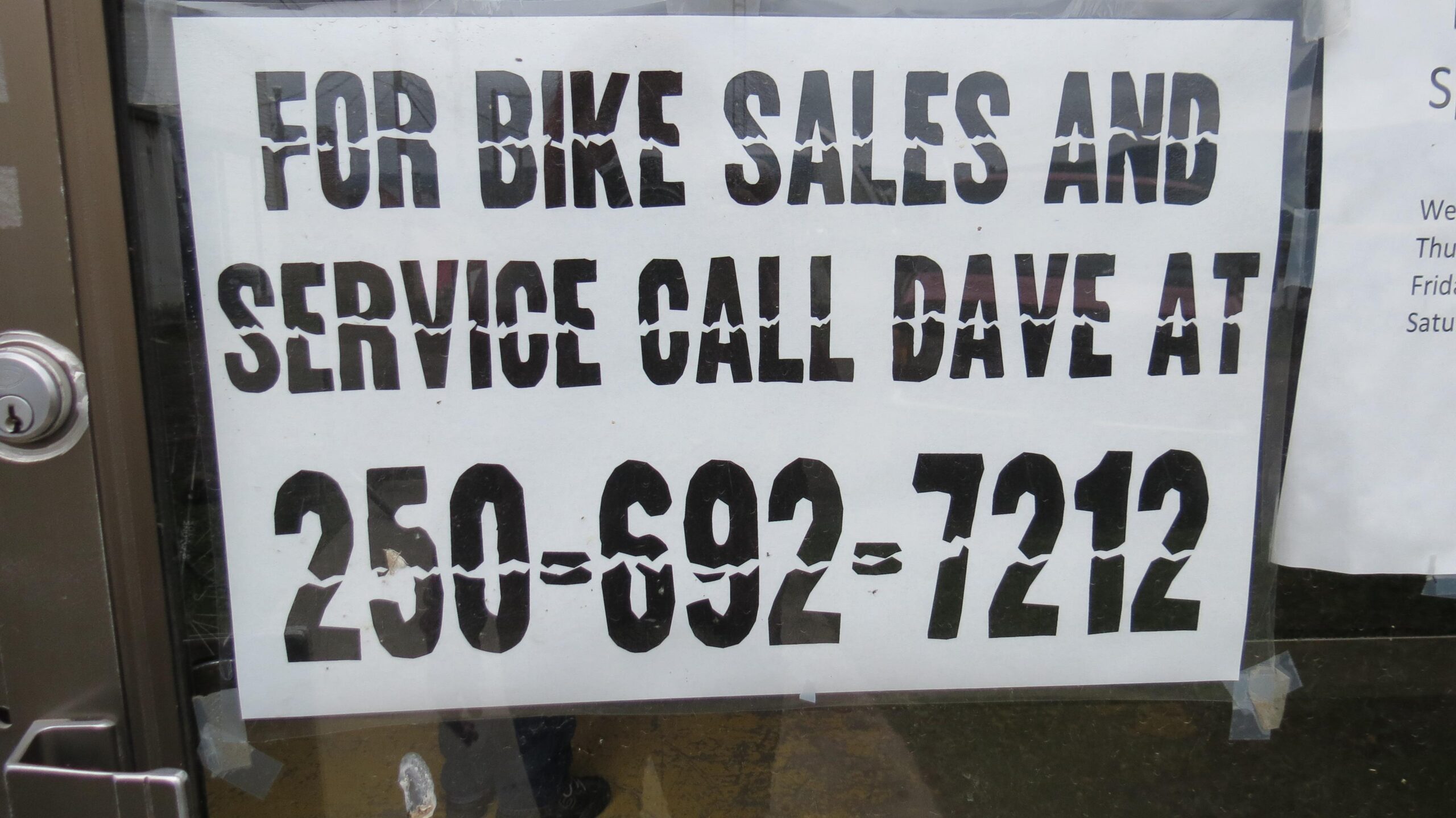 A printed sign on a storefront window advertising bike sales and service, instructing customers to call Dave at the phone number 250-692-7212. The sign features bold black lettering on a white background.