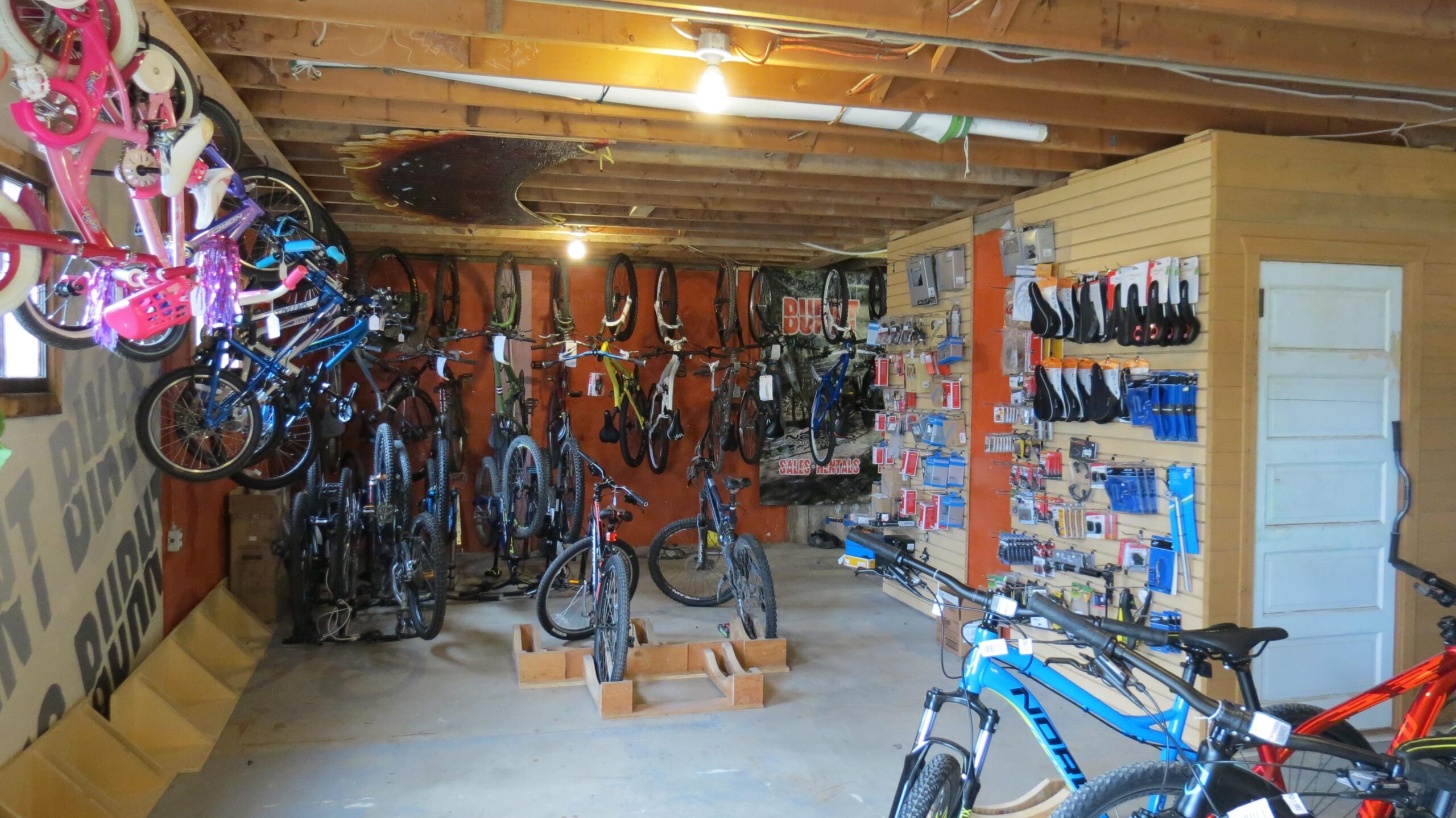 A spacious bike shop interior featuring a variety of bicycles, including children's bikes in bright colors hanging from the walls. The shop displays several mountain and road bikes on the floor and walls, along with bicycle accessories and tools organized on shelves. A wooden structure in the center serves as a display area for additional bikes. The ceiling is exposed with overhead lighting illuminating the space.