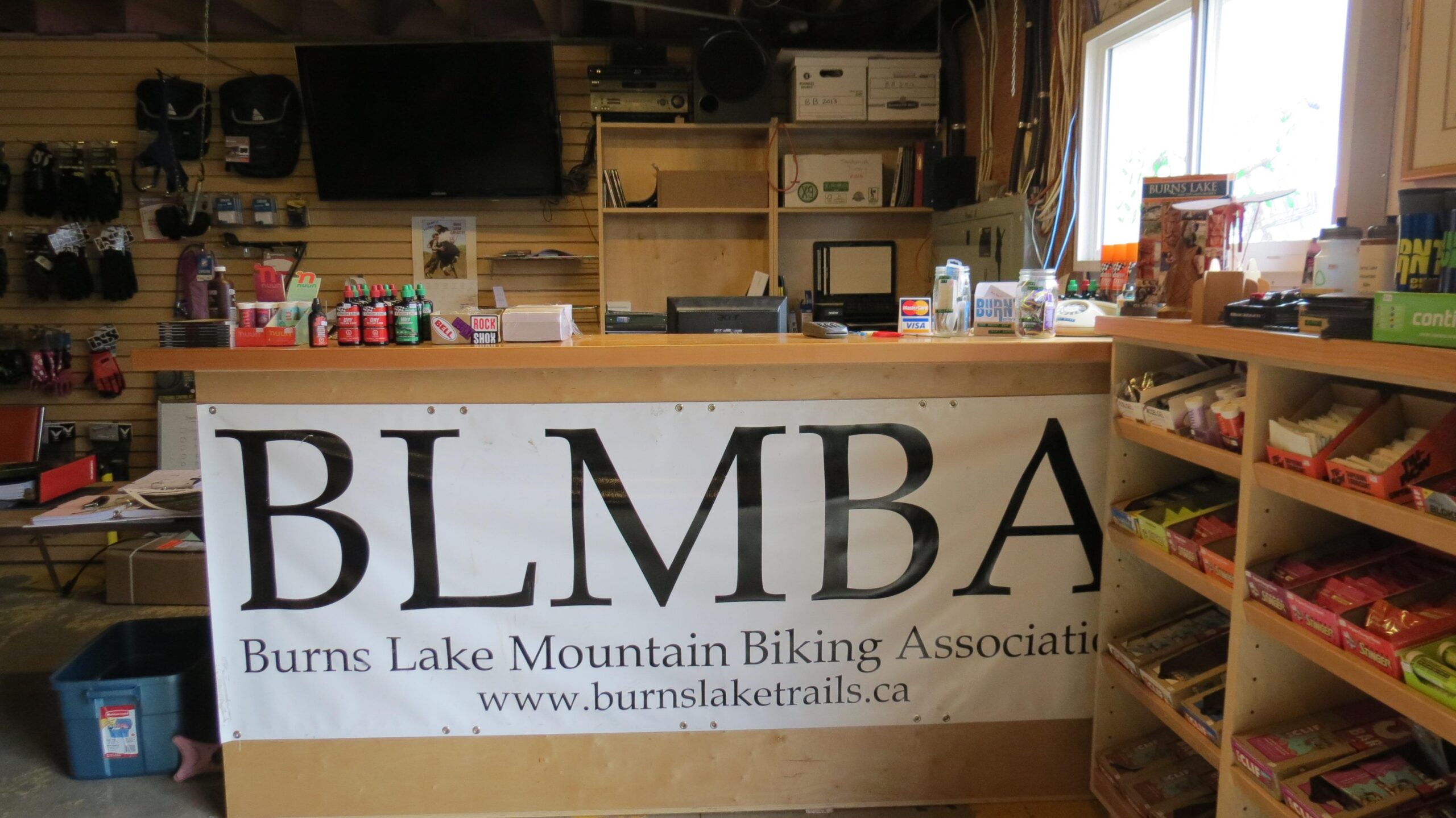 Interior view of a shop featuring a wooden counter with a banner displaying "BLMBA - Burns Lake Mountain Biking Association." The background includes shelves stocked with biking gear, snacks, and beverages, along with a television and various office supplies.