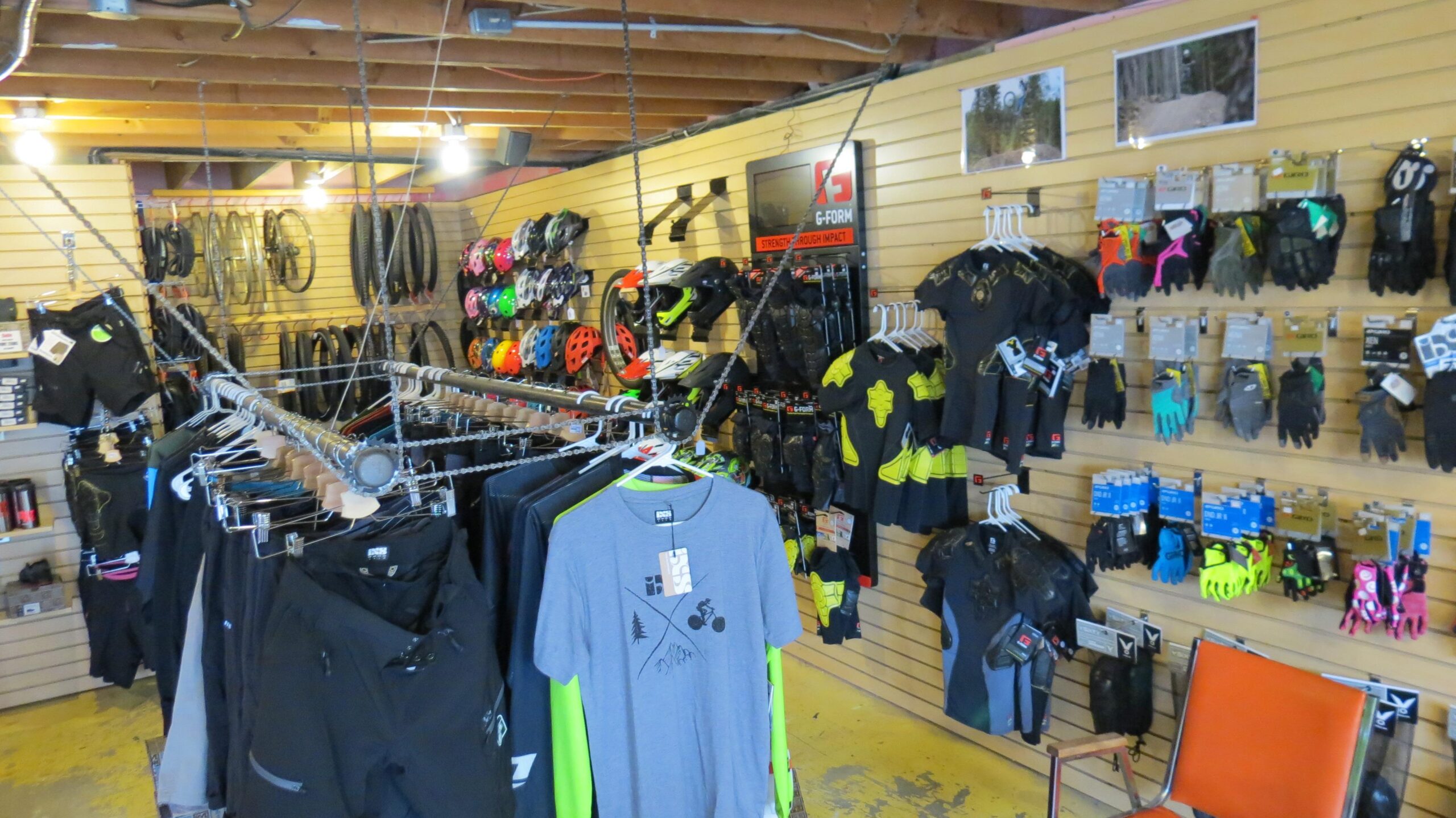 A retail store interior featuring a variety of cycling gear and apparel. The walls are lined with helmets, gloves, and protective clothing, while racks display various shorts and shirts. The space has wooden beams and is brightly lit, creating a welcoming atmosphere for customers interested in biking equipment.