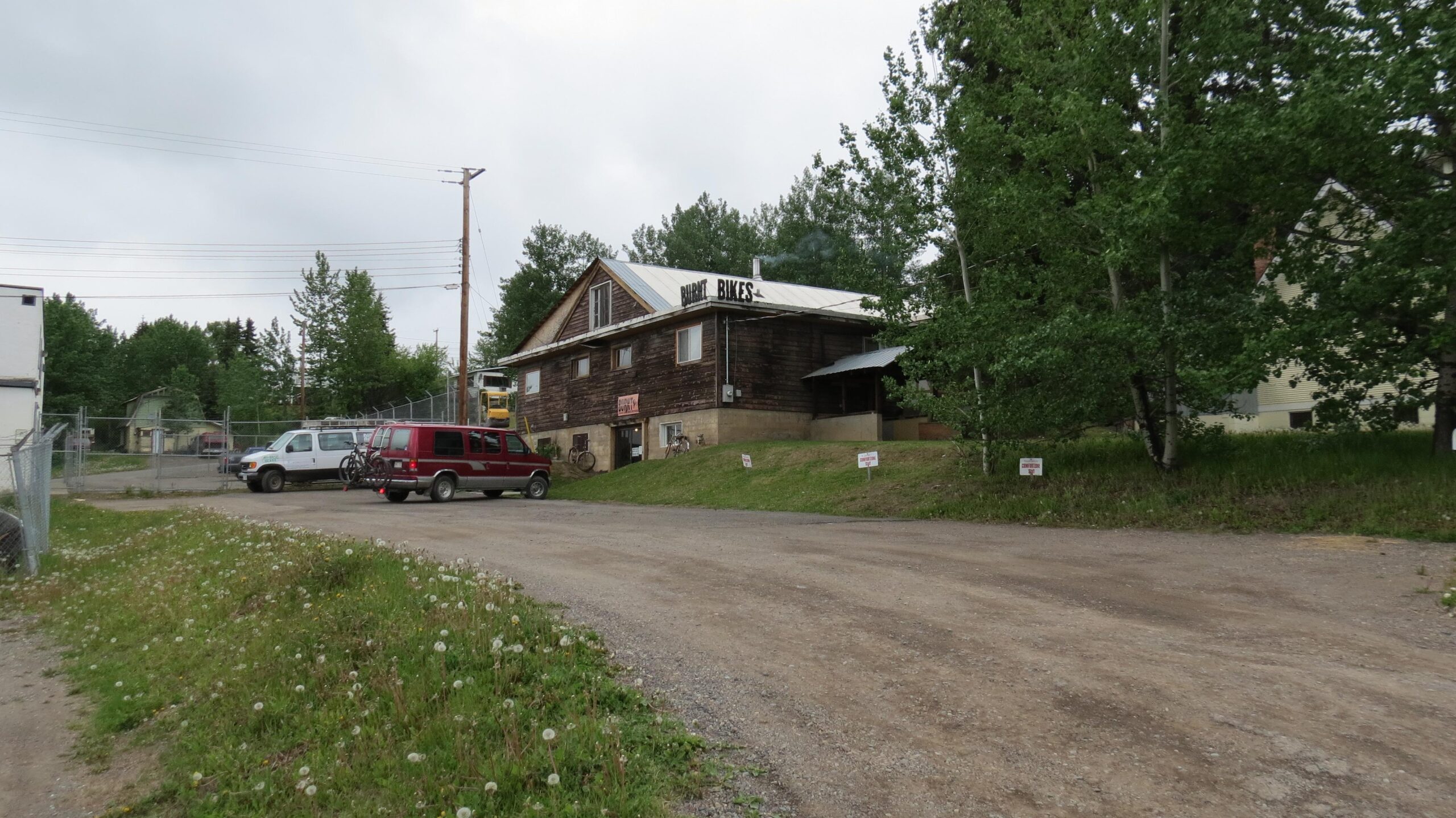A gravel road leads to a rustic wooden building with a sign that reads "Burt Bikes." Several vehicles, including a red van and a white pickup truck, are parked in front of the building. Surrounding the area are green trees and tall grass, with dandelions growing in the foreground. The sky is cloudy, suggesting a typical overcast day.