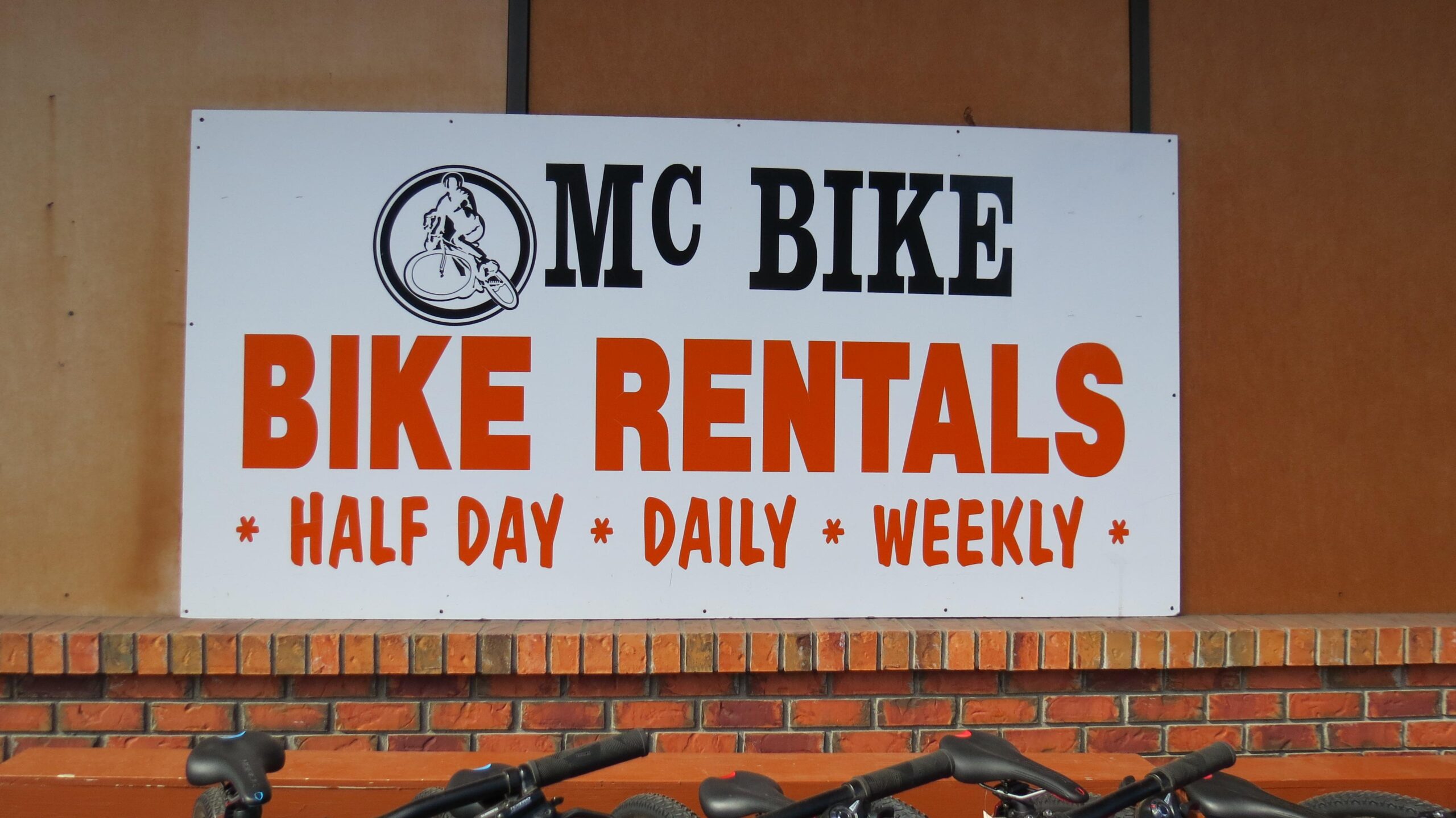 Sign for "MC Bike" showing rental options for bicycles, including half-day, daily, and weekly rentals, with a brick wall in the background and bike handlebars visible in the foreground.