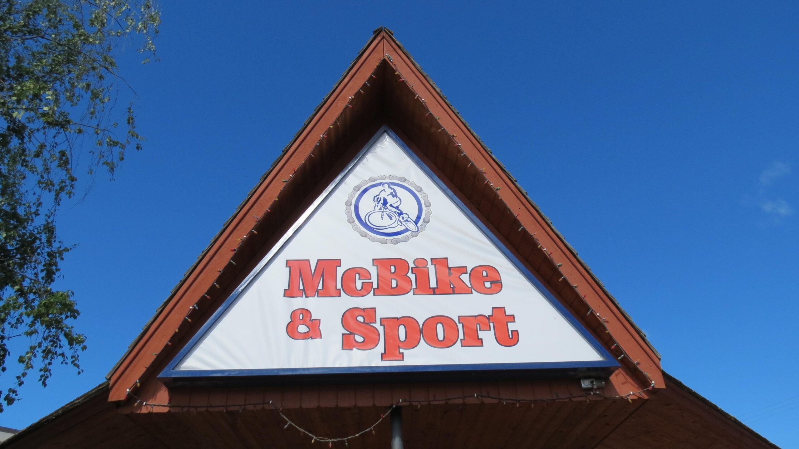 Sign for McBike & Sport, featuring a cyclist graphic, against a clear blue sky. The sign is mounted on a triangular wooden structure with decorative lights.