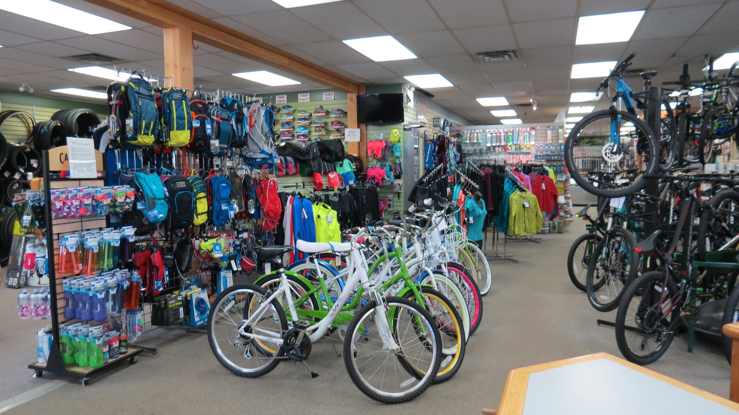Interior view of a bike shop featuring various bicycles in the foreground, including colorful models with white, green, blue, and pink accents. The background showcases shelves stocked with backpacks, sports gear, and accessories, including hydration bottles and clothing. The shop is well-lit with a variety of products displayed on racks and walls.