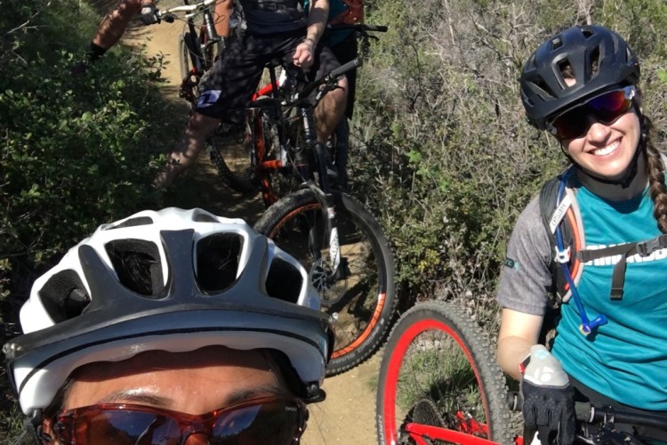 A group of four mountain bikers taking a selfie on a sunny trail, surrounded by greenery and hills. One cyclist is making a playful face in the foreground, while others can be seen smiling and giving thumbs up in the background. The cyclists are wearing helmets and sunglasses, with their bikes parked alongside the trail.