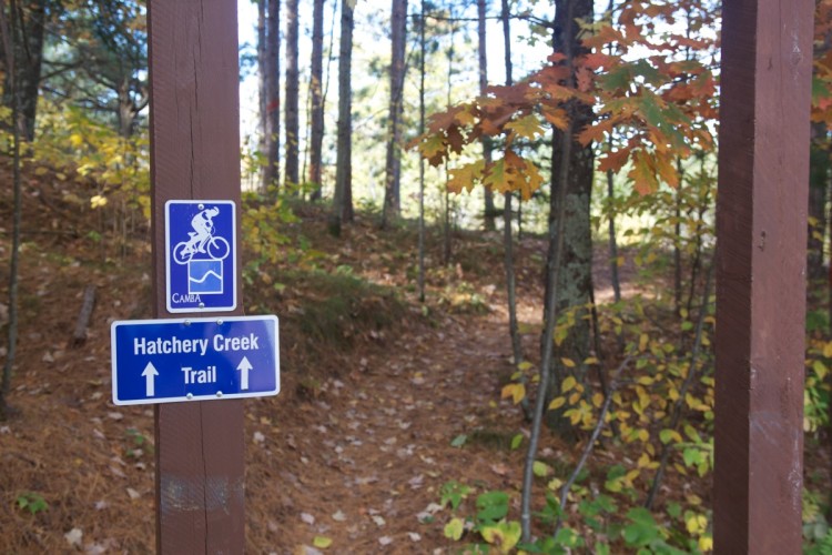 A wooden trail sign indicating the Hatchery Creek Trail, with a bike symbol above it. The background features a wooded area with trees showing autumn foliage.